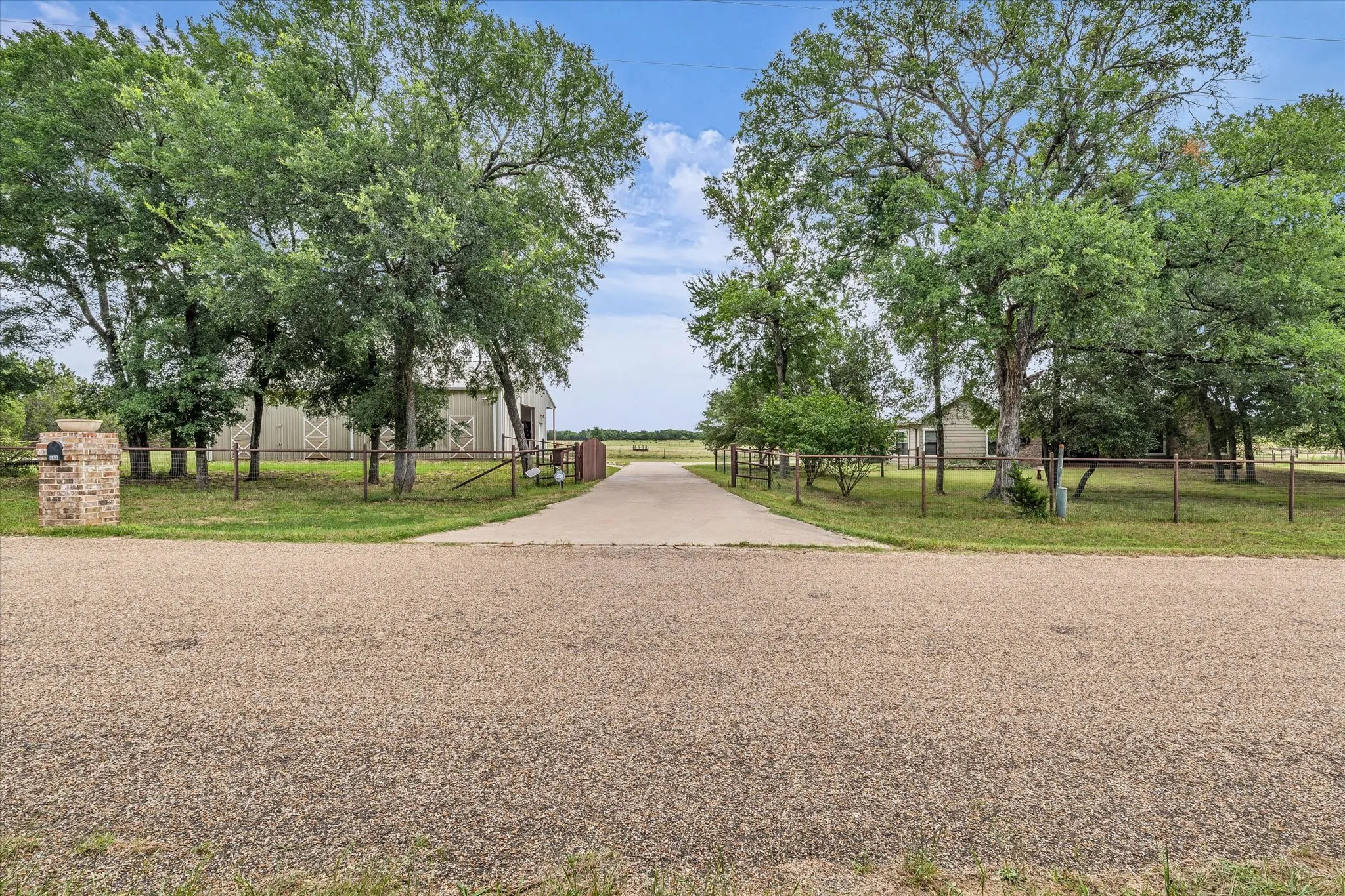 Paved driveway entry framed by mature trees, leading to a property featuring a brick mailbox column, perimeter fencing, and a barn structure