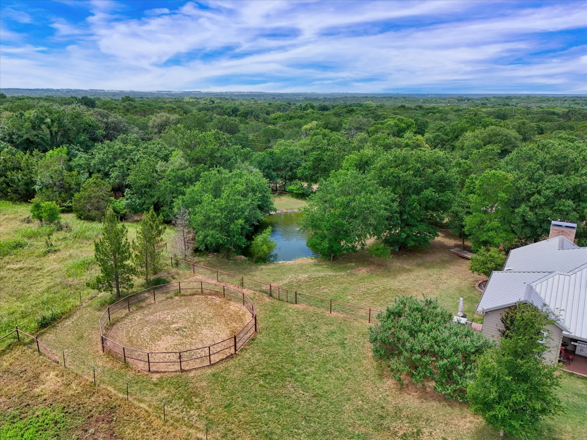Expansive property featuring a tranquil pond surrounded by mature trees, a fenced round pen, and a residence with a metal roof and brick chimney