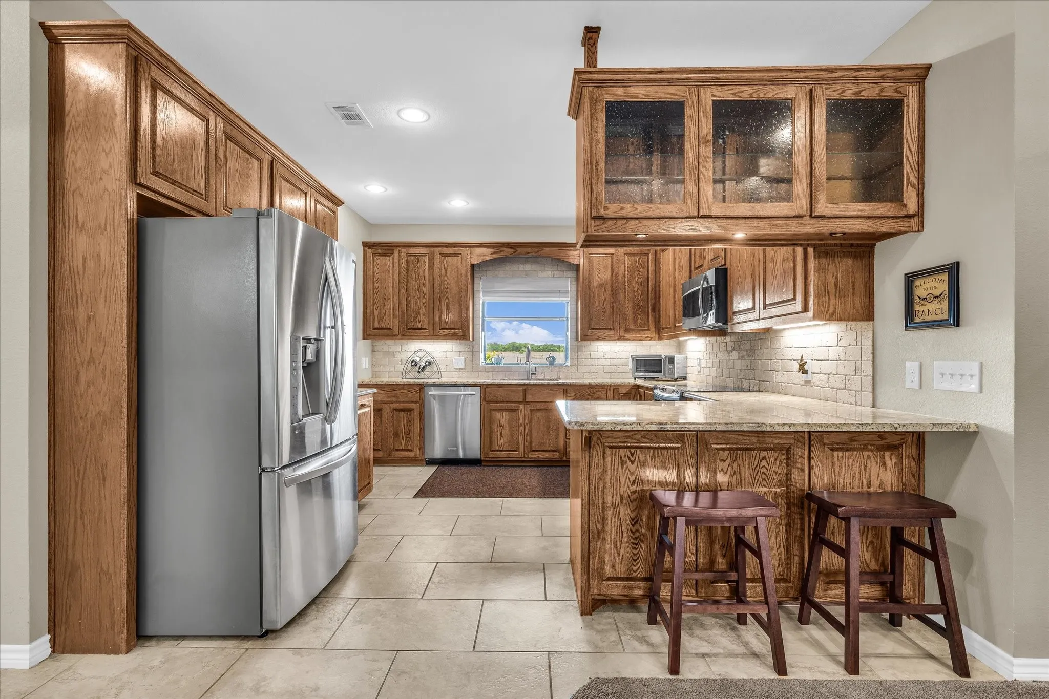 Kitchen featuring wood cabinetry, light-toned tile flooring, recessed lighting, a window over the sink, and a breakfast bar with stone countertops