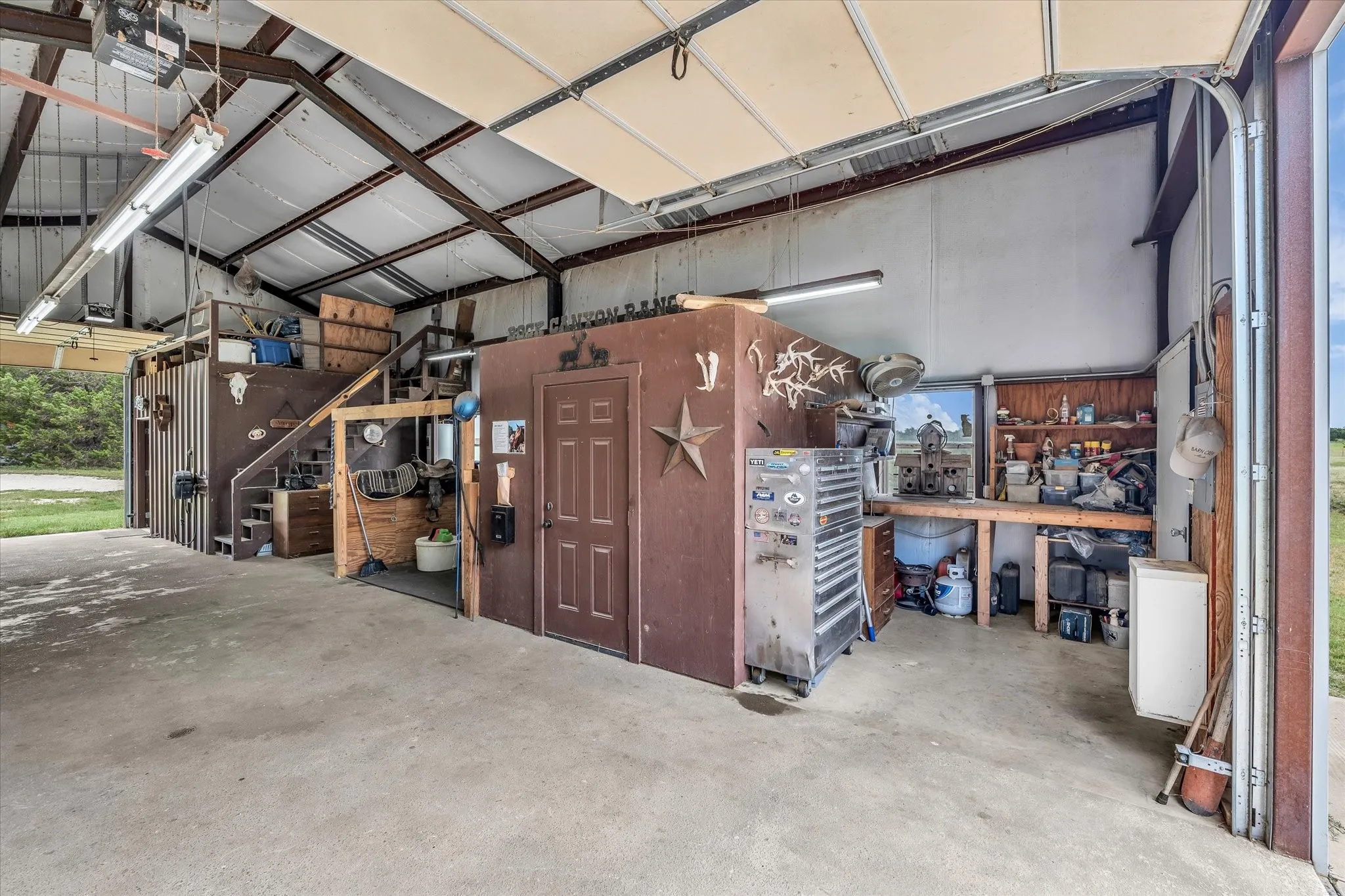 Spacious utility building featuring a concrete floor, exposed metal beam structure, multiple fluorescent light fixtures, a roll-up garage door, and a built-in workbench with shelving