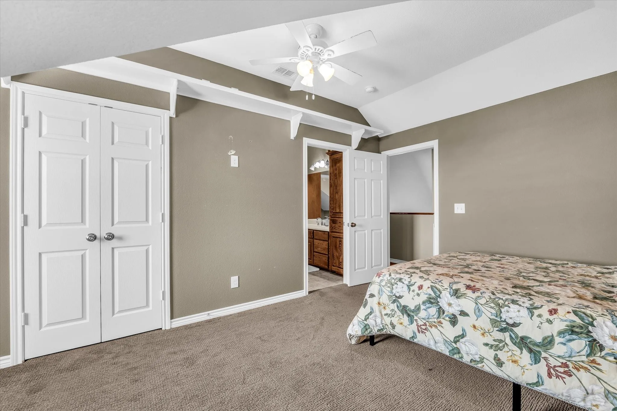 Carpeted room featuring a vaulted ceiling with a white ceiling fan, white bi-fold closet doors, and direct access to an attached bathroom with a wood-finish vanity