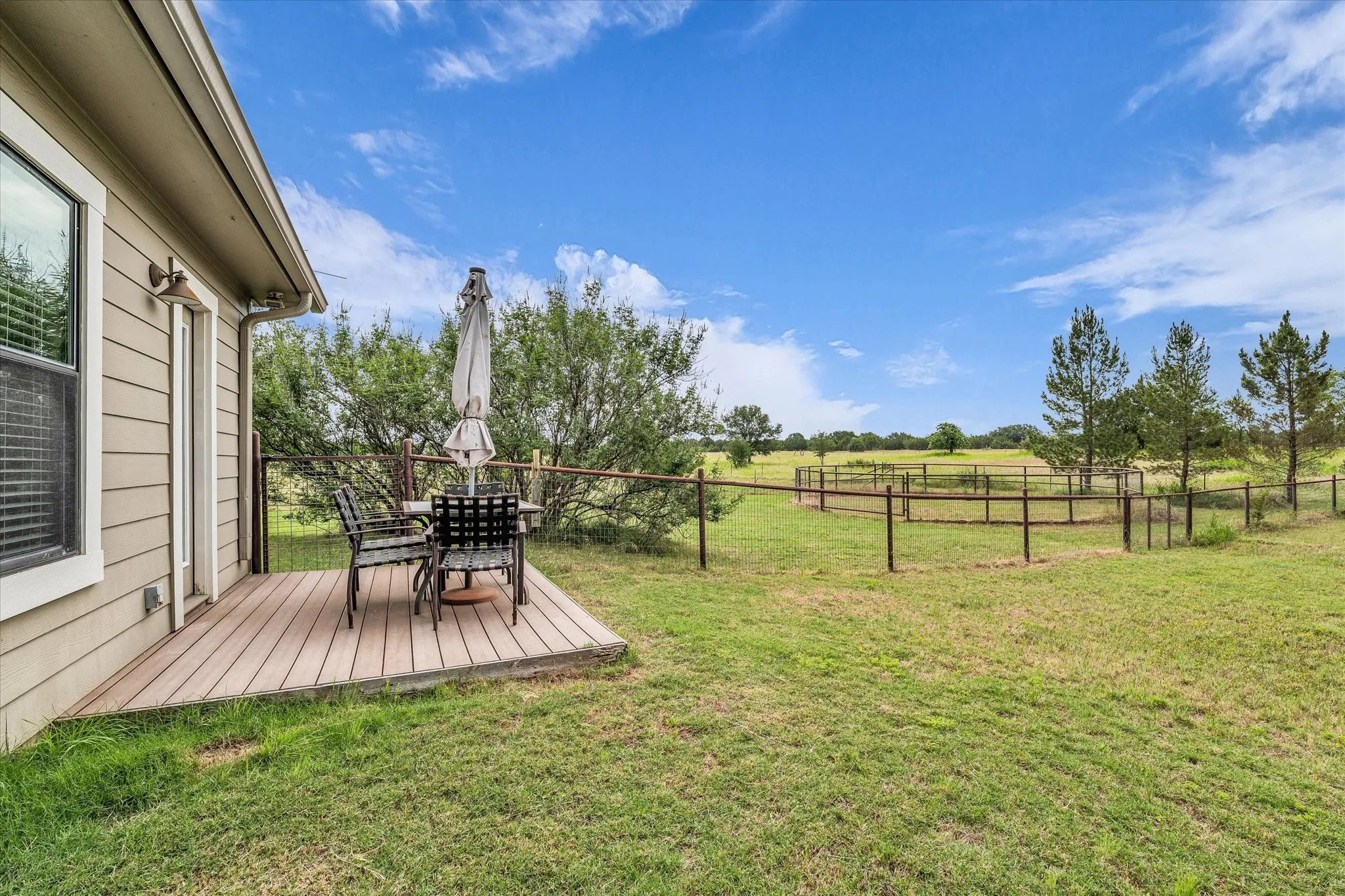 Outdoor wooden deck with siding exterior, featuring a fenced pasture beyond the property line, mature trees, and an expansive lawn