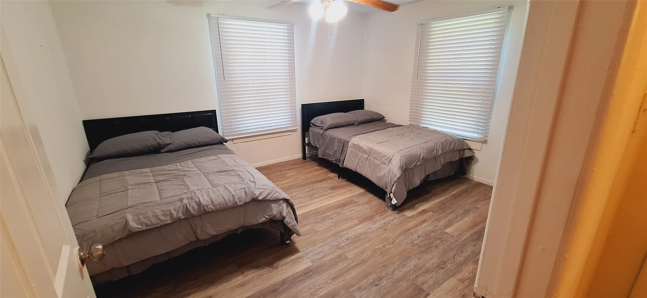 Room featuring wood-finish flooring, two large windows with blinds, a ceiling fan with integrated lighting, and white trim