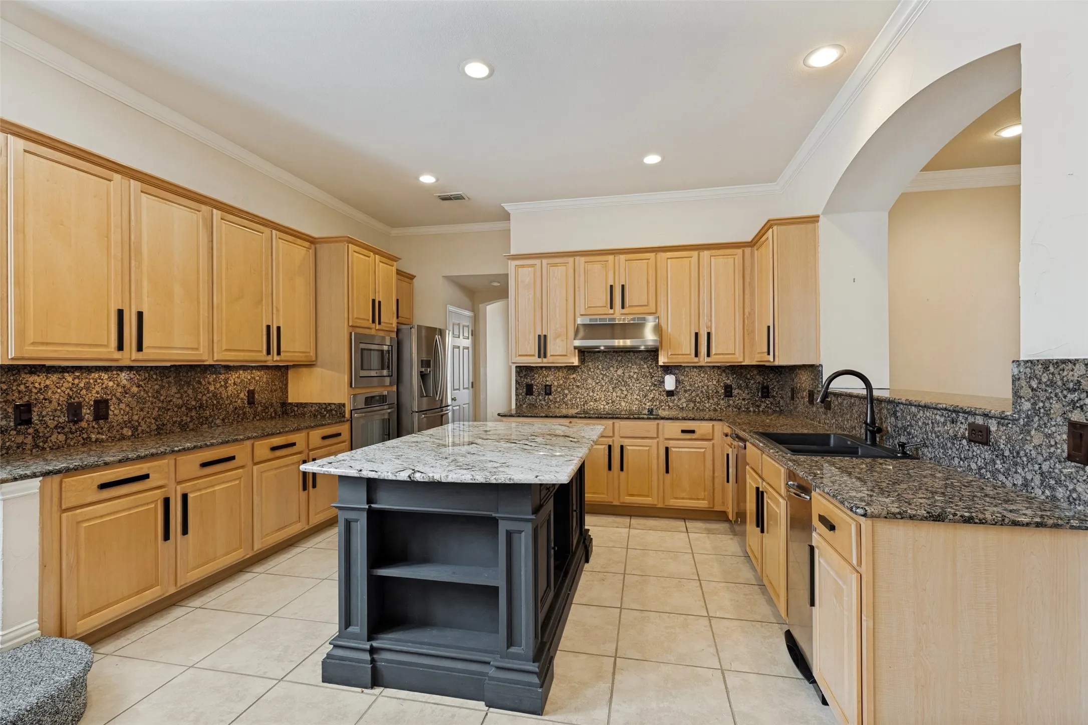 Kitchen featuring a central island with a contrasting dark base and light-colored countertop, extensive light wood cabinetry with dark hardware, speckled stone countertops and backsplash, stainless steel appliances, and recessed lighting