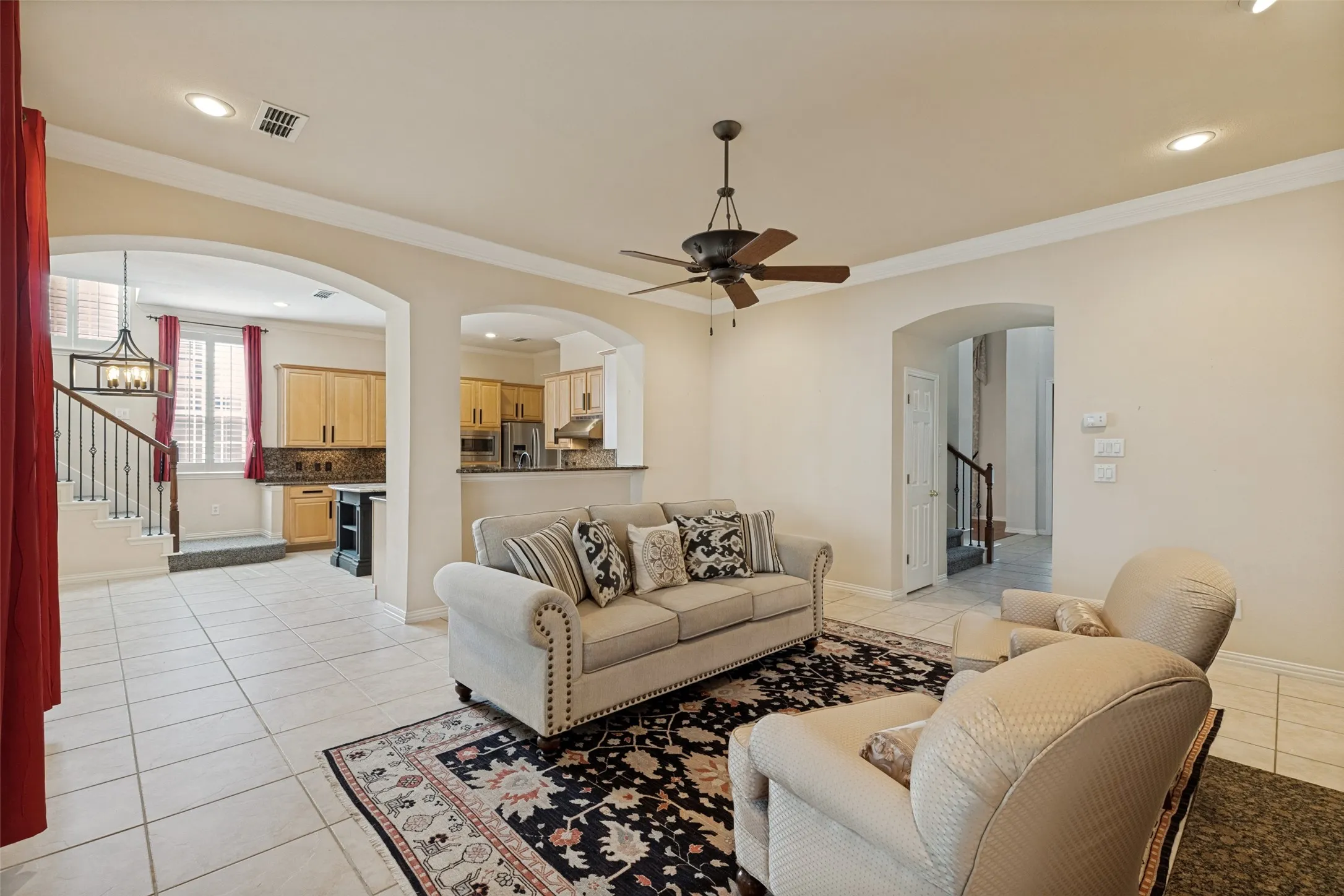 Arched doorways framing a bright interior with tile flooring throughout, a ceiling fan with light fixture, recessed lighting, and a view into the kitchen featuring light wood cabinetry