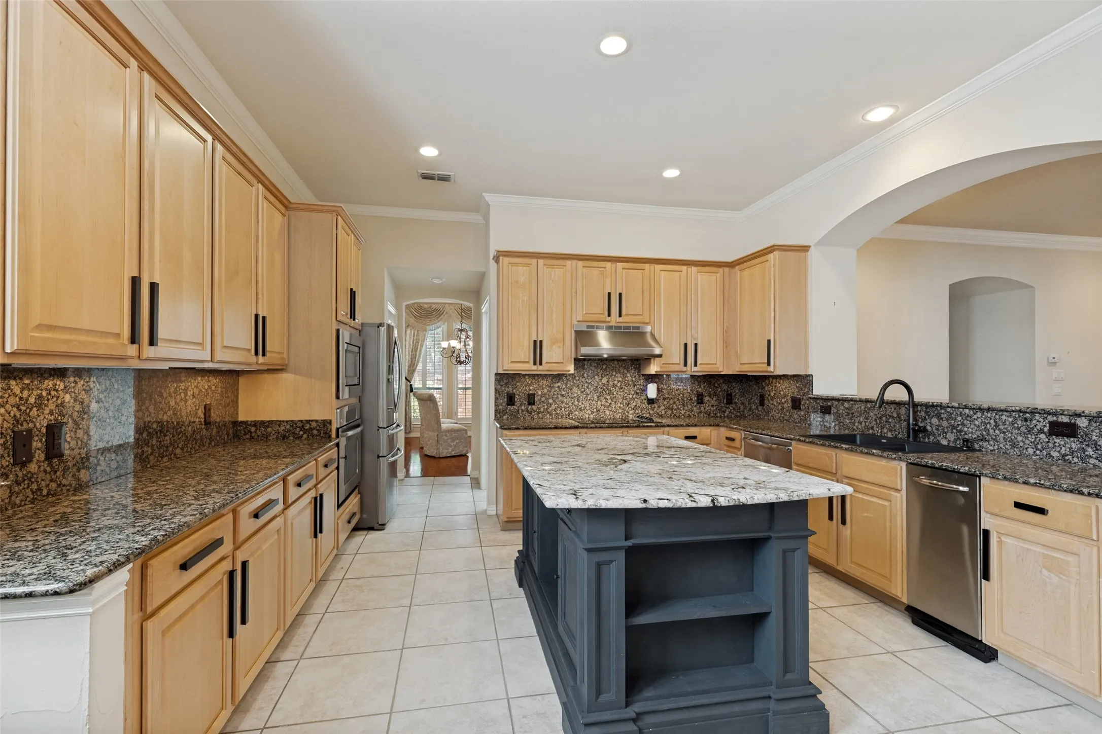 Kitchen featuring light wood cabinetry with black hardware, granite countertops, tile flooring, a central island with open shelving, and stainless steel appliances