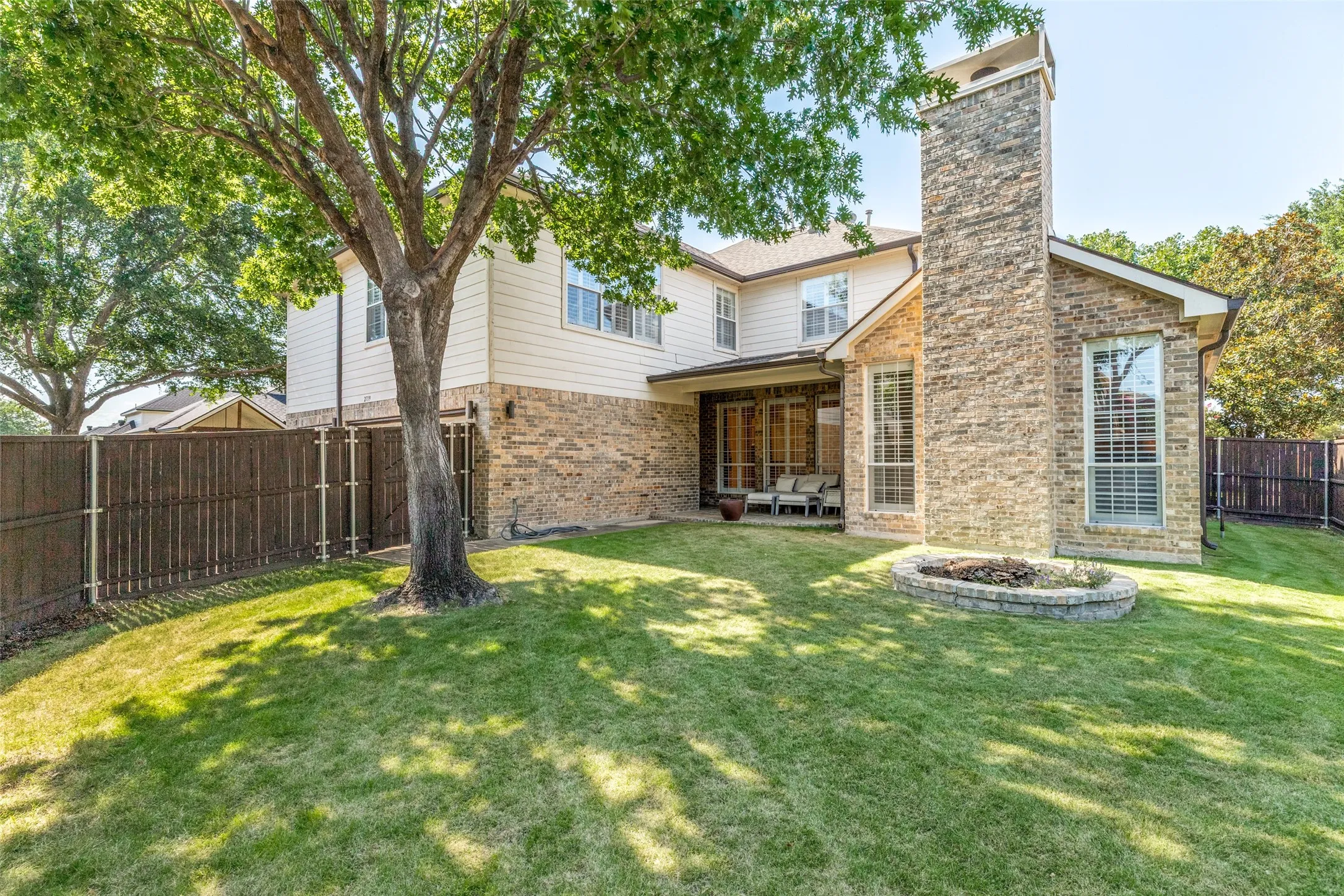 Expansive backyard featuring a manicured lawn, a mature shade tree, and a stone-clad exterior with a prominent chimney
