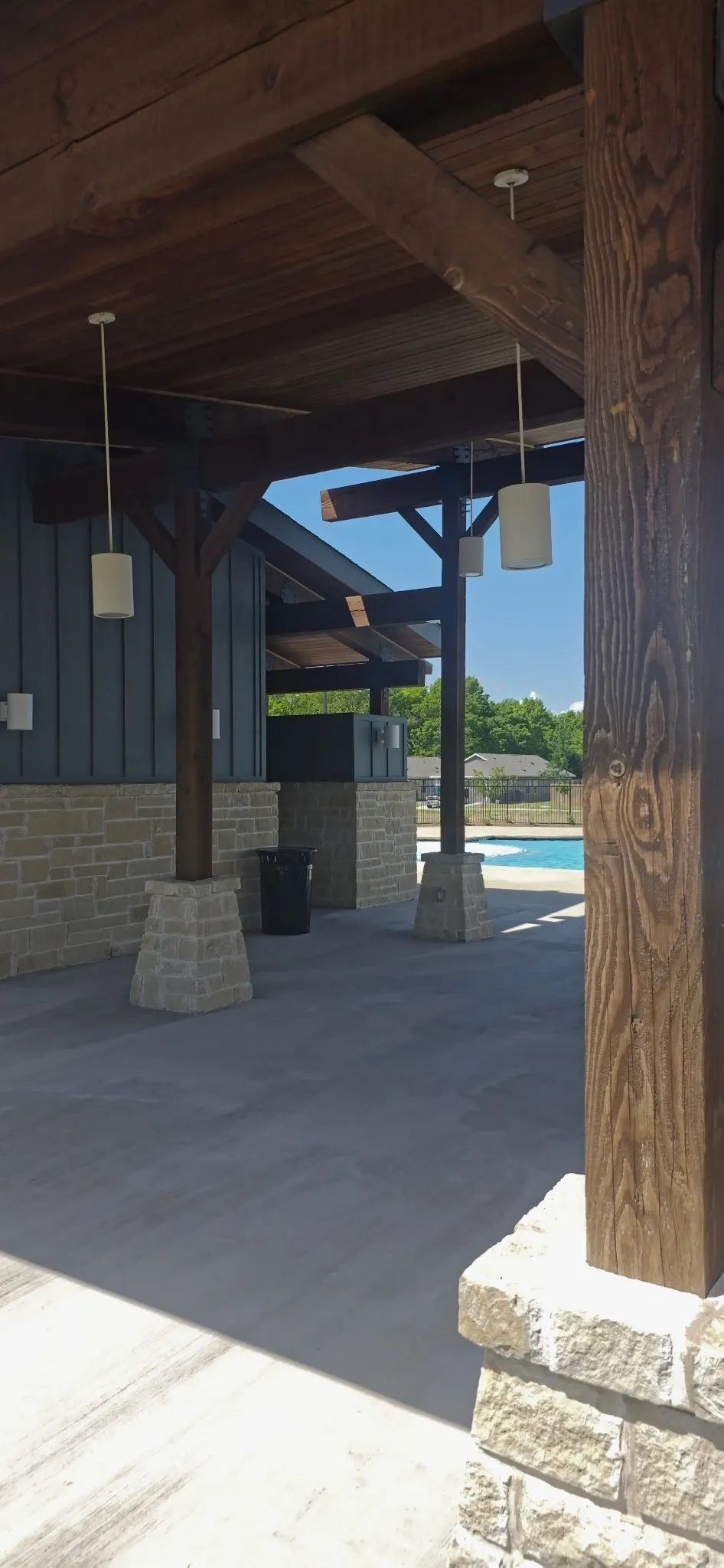 Covered outdoor amenity space featuring exposed wood beams, stone column bases, pendant lighting, and a concrete floor