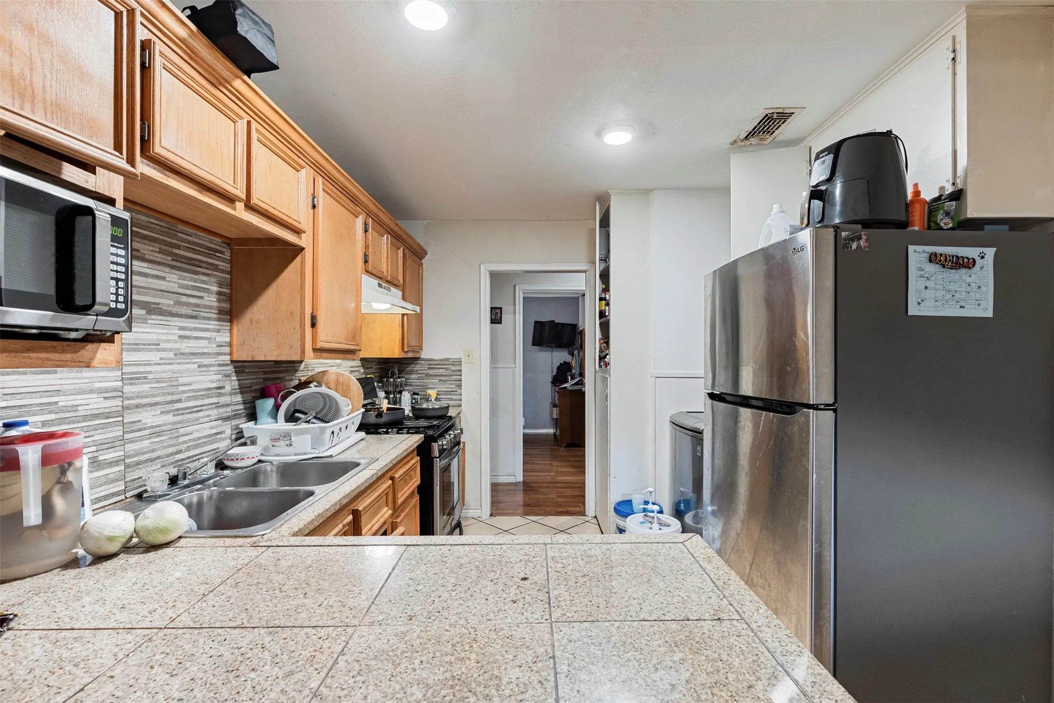 Kitchen featuring wood cabinetry, a tiled countertop, a mosaic tile backsplash, a double basin sink, and a stainless steel refrigerator