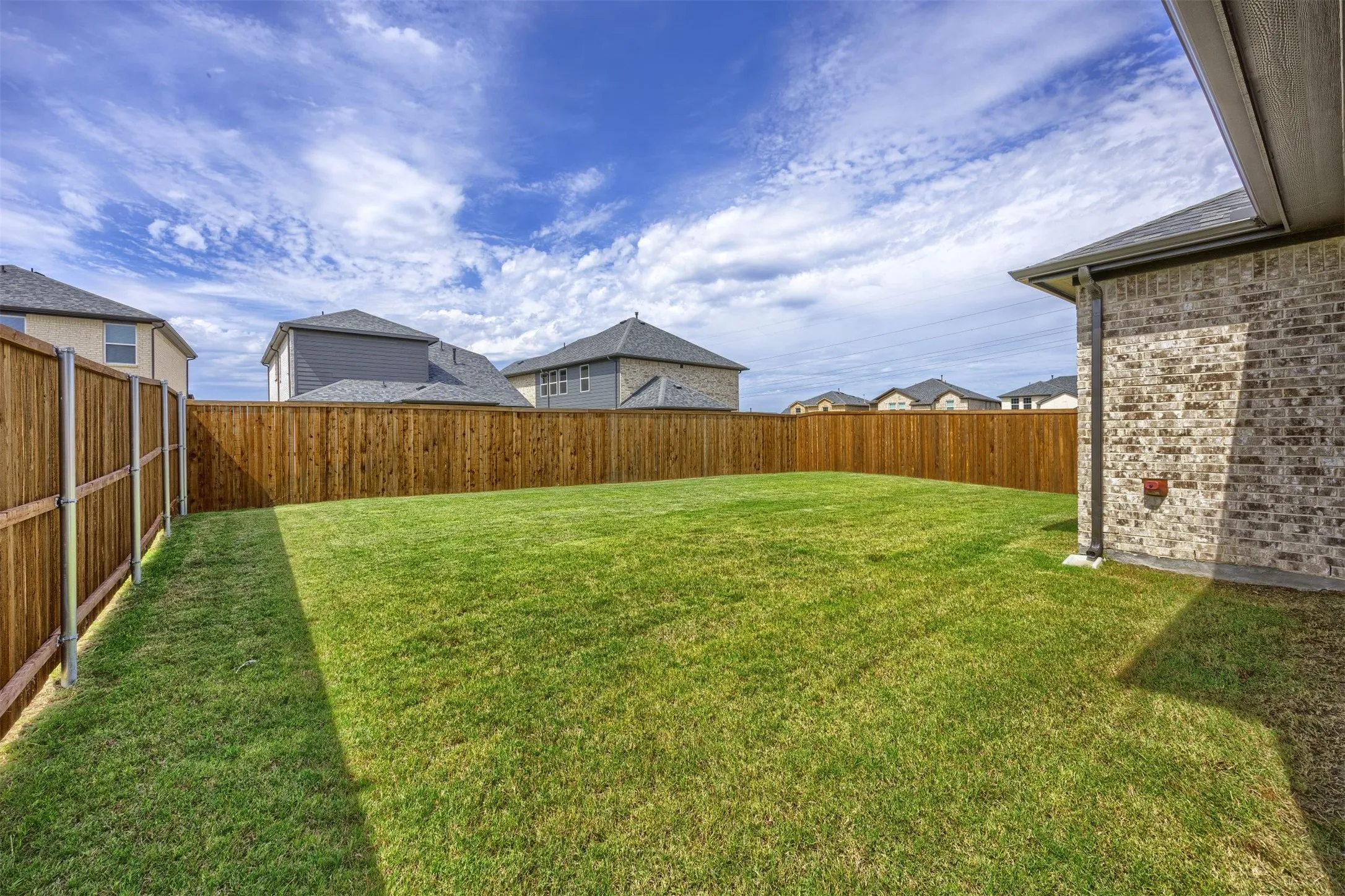 Expansive grassy backyard featuring a full privacy fence, brick exterior siding, and a partial covered patio area