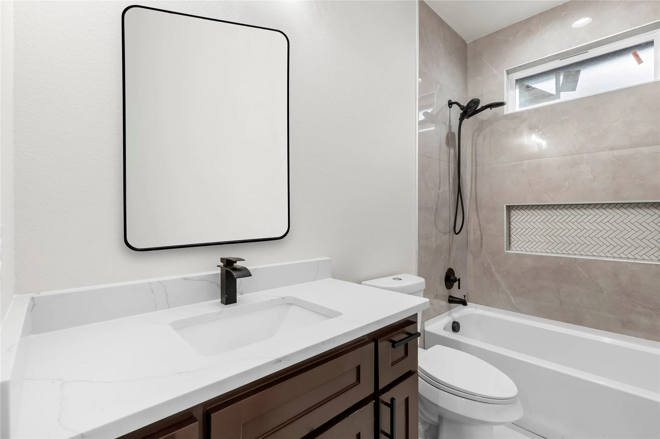 Bathroom featuring a dark wood-finish vanity with a white countertop and integrated sink, matte black fixtures, a rectangular framed mirror, and a bathtub with full-height tile surround and a tiled niche