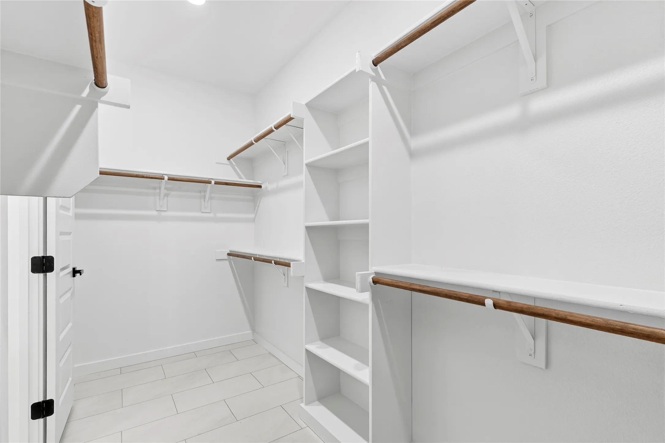Walk-in closet featuring white built-in shelving, multiple hanging rods, light-toned tile flooring, and a white paneled door with black hardware