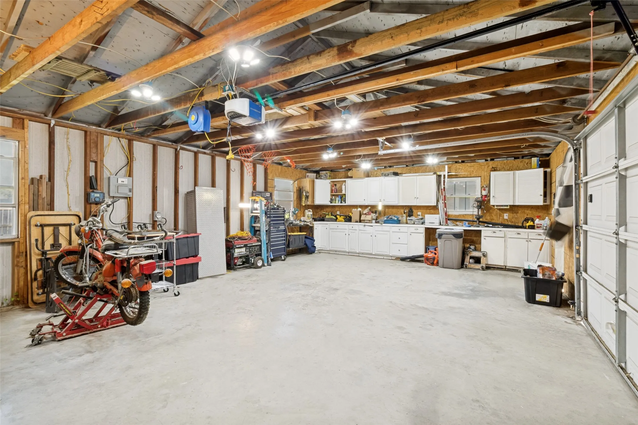 Spacious garage featuring exposed wood beam ceilings, extensive built-in cabinetry, durable concrete flooring, and multiple overhead light fixtures