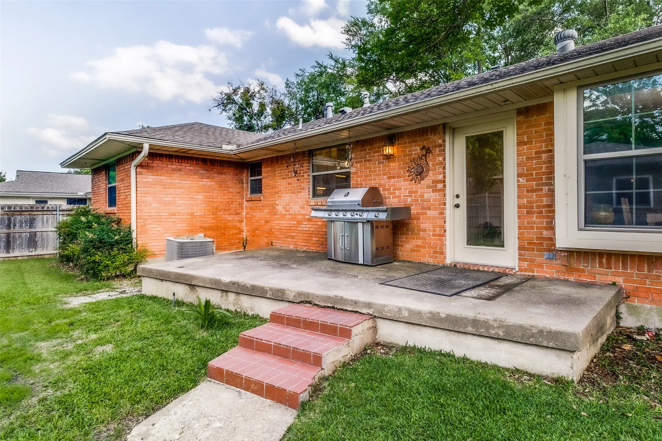 Back of house featuring a concrete patio with brick steps, a back door, and windows overlooking a grassy area