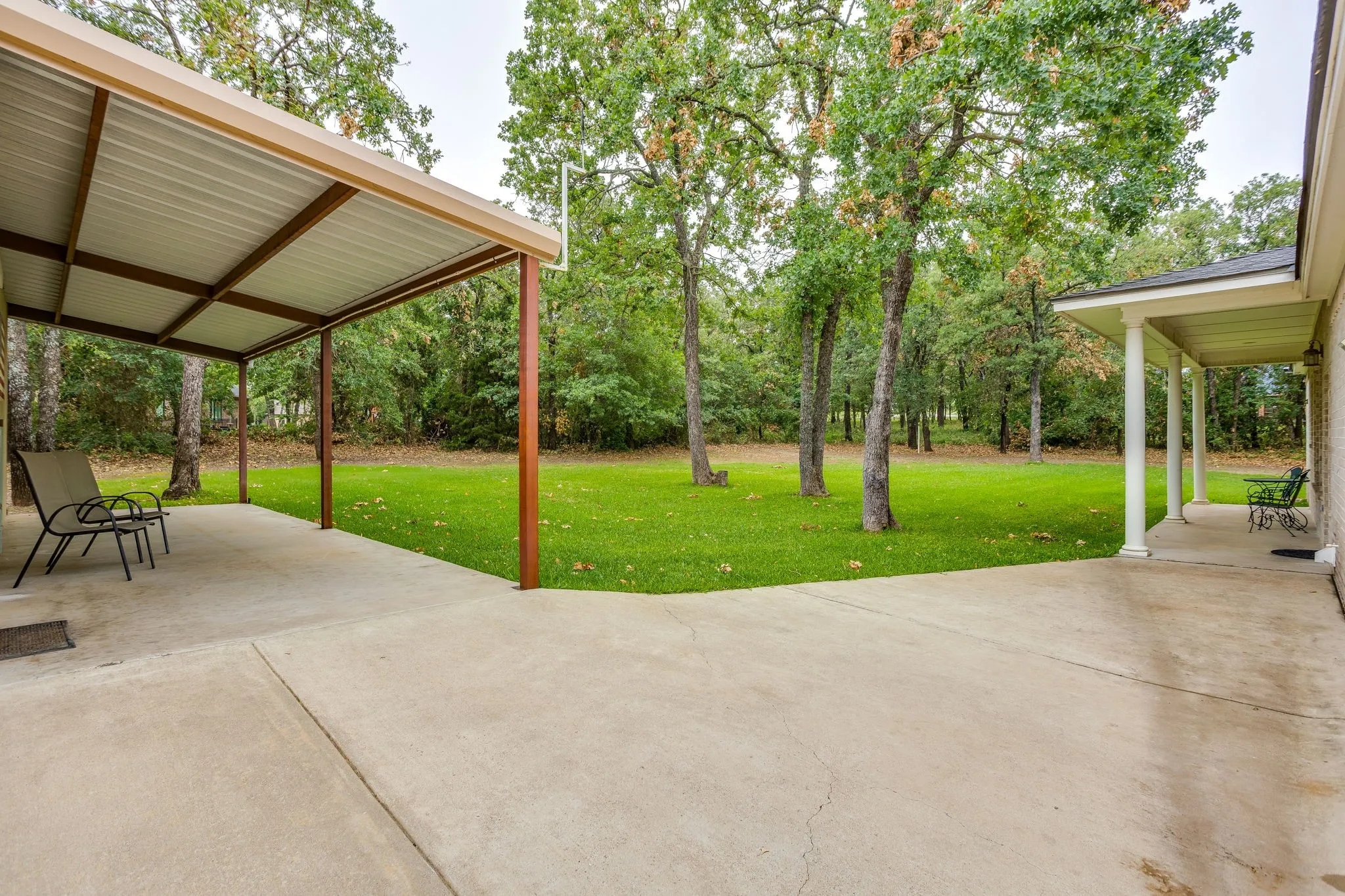 Expansive concrete patio area featuring a covered carport structure with a metal roof, a second covered porch with white columns, a large grassy yard, and mature trees