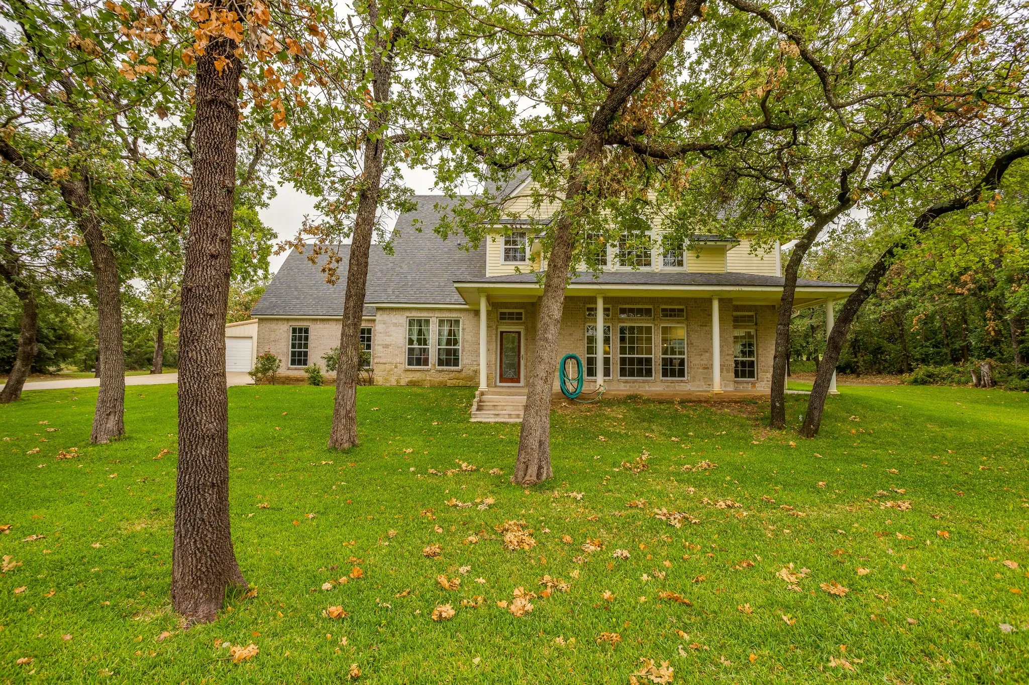 Spacious lawn with mature trees, stone and siding exterior, multiple windows, covered porch, and an attached garage