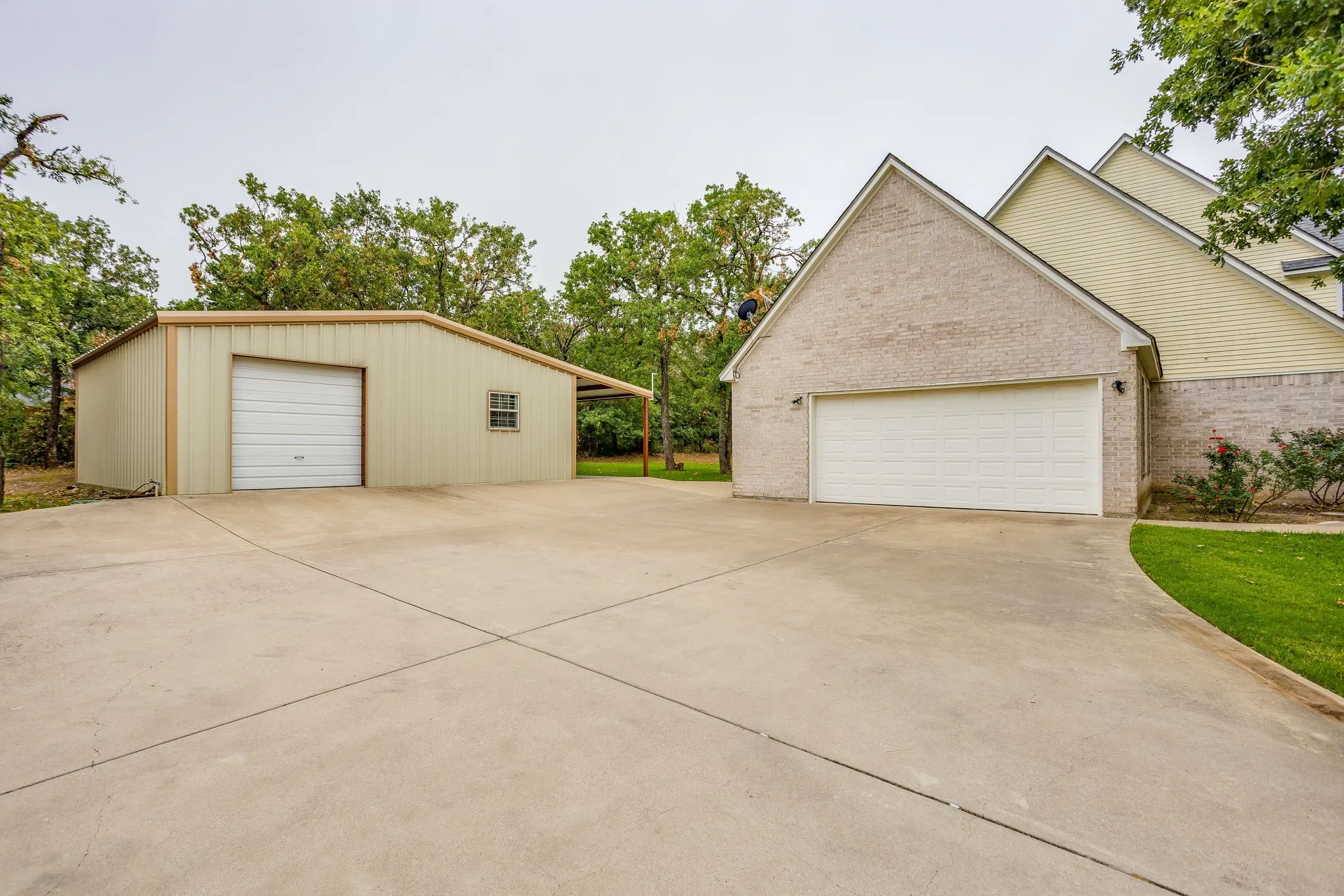 Expansive concrete driveway leading to a multi-car attached garage and a detached metal workshop with a single garage door and covered carport