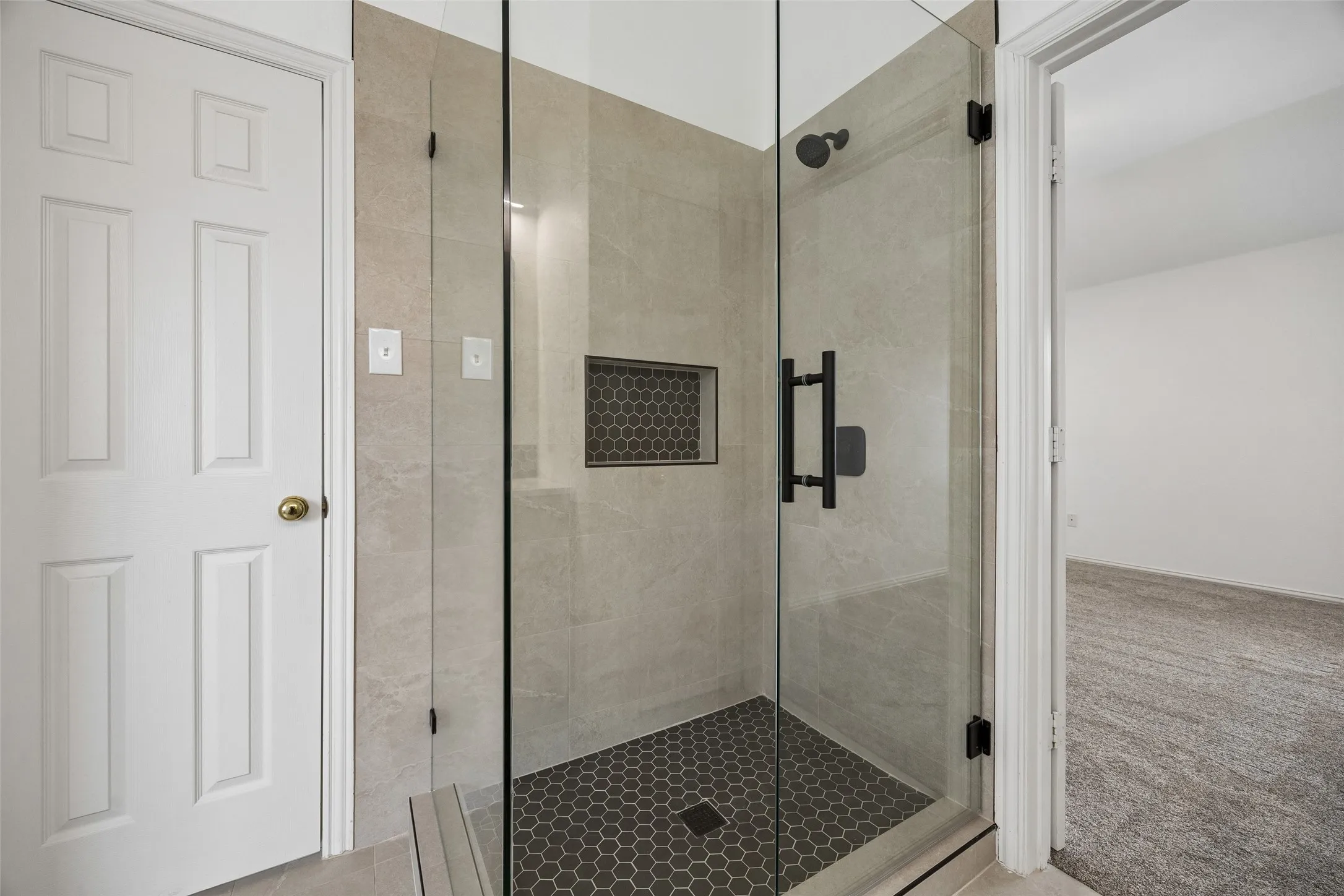 Contemporary walk-in shower featuring clear glass enclosure, matte black fixtures, light-toned stone-finish wall tiling, dark hexagonal floor tiling, and a recessed hexagonal tile niche