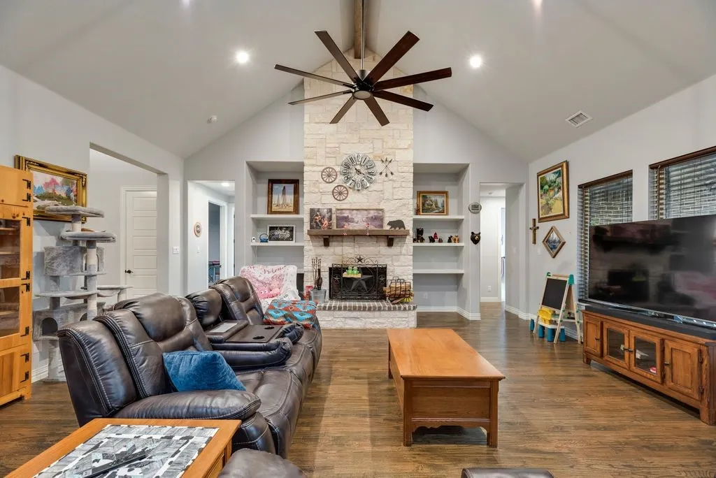 Spacious living area featuring a vaulted ceiling with a ceiling fan, a stone-faced fireplace, and wood-finish flooring
