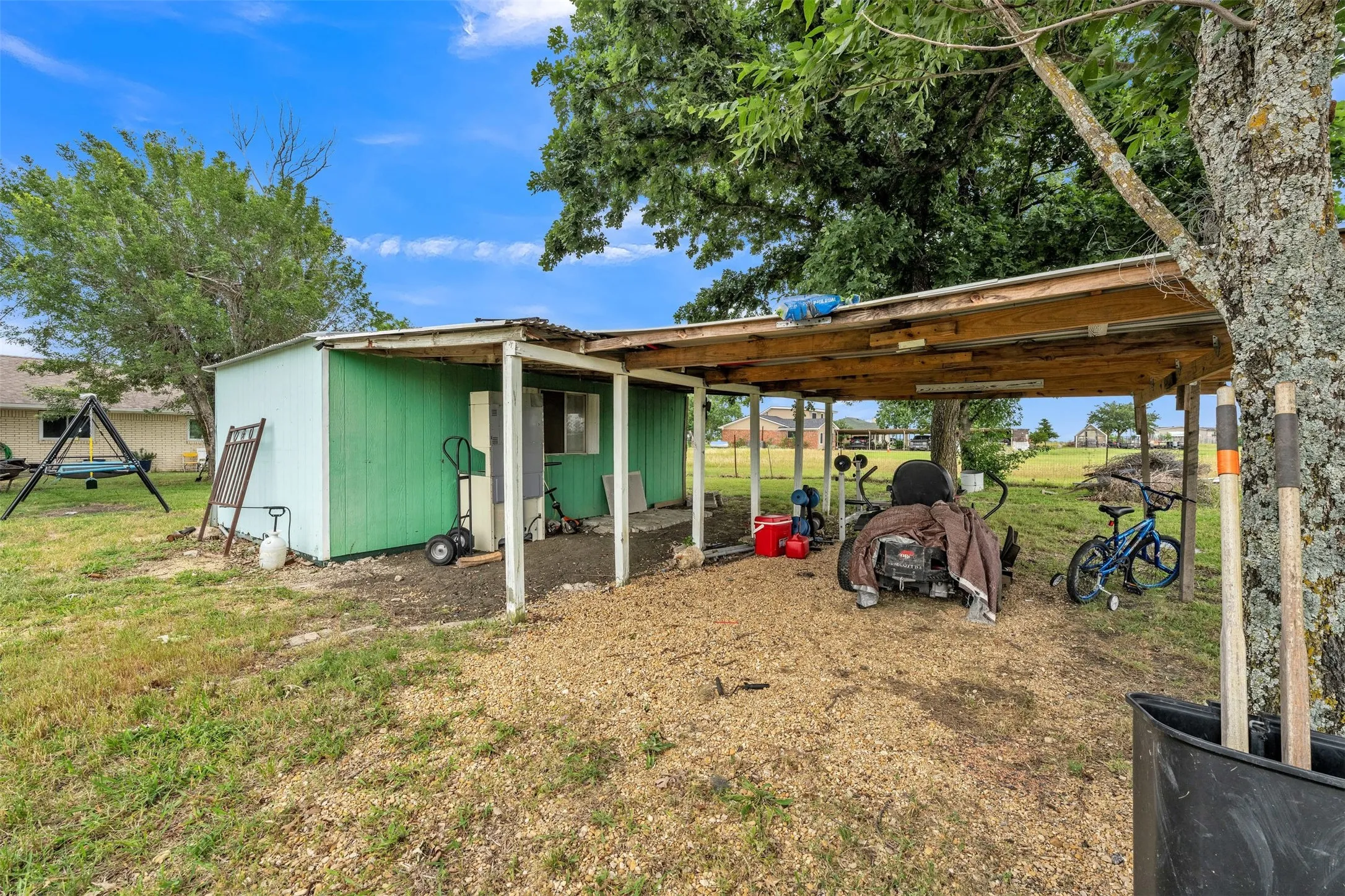 Detached utility structure with green siding and an attached covered carport featuring a wood-frame roof and gravel flooring