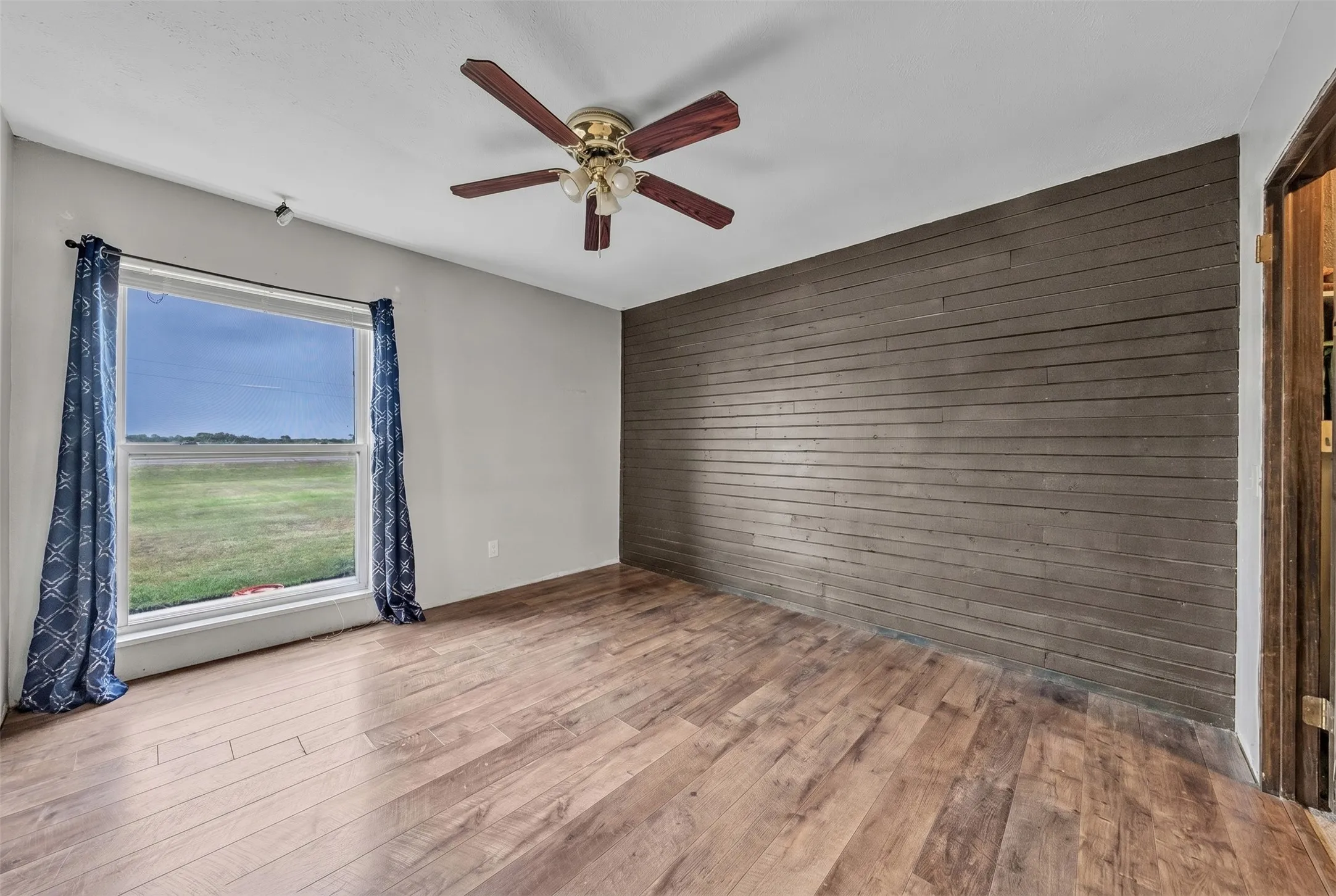 Spacious room featuring wood-finish flooring, a decorative wall panel, and a ceiling fan with integrated lighting