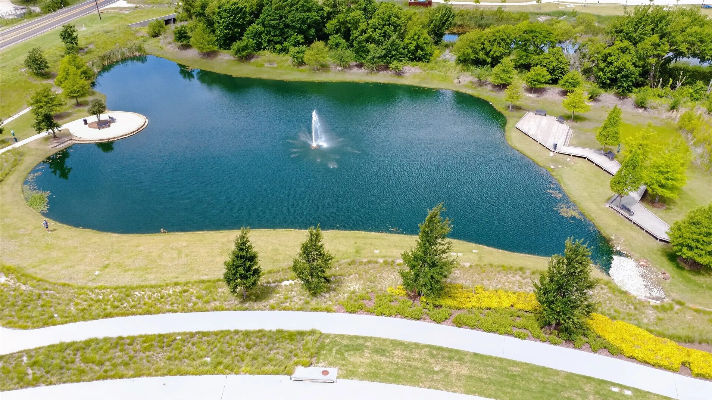 Expansive water feature with a central fountain, bordered by a walking path, landscaped grounds, and a wooden boardwalk with seating areas