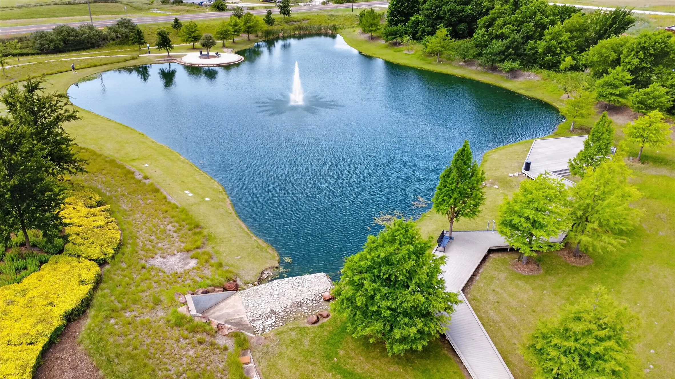 Expansive pond featuring a central fountain, an island with seating, a concrete walkway, and surrounding green spaces with mature trees and landscaped shrubbery