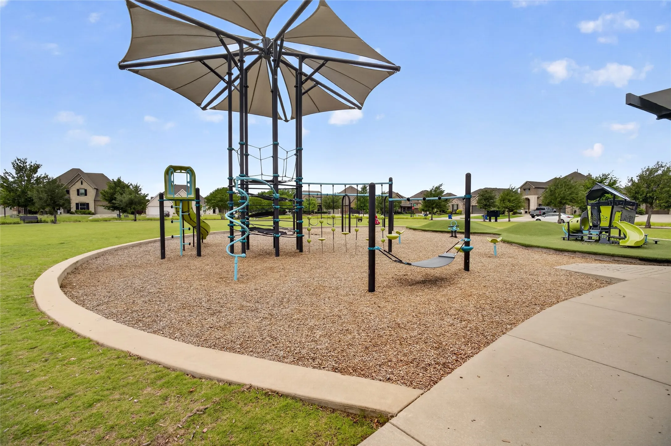 Community playground featuring a shade structure, multiple play apparatuses, wood chip surfacing, and a concrete pathway