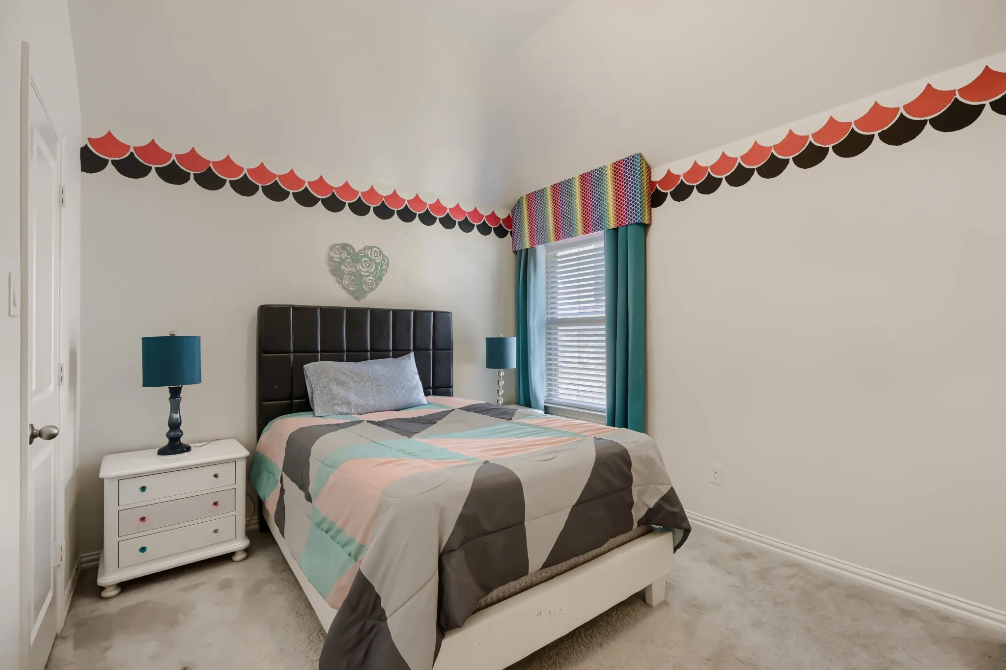 Carpeted bedroom featuring an upholstered headboard, white trim, and a window with blinds and colorful valance