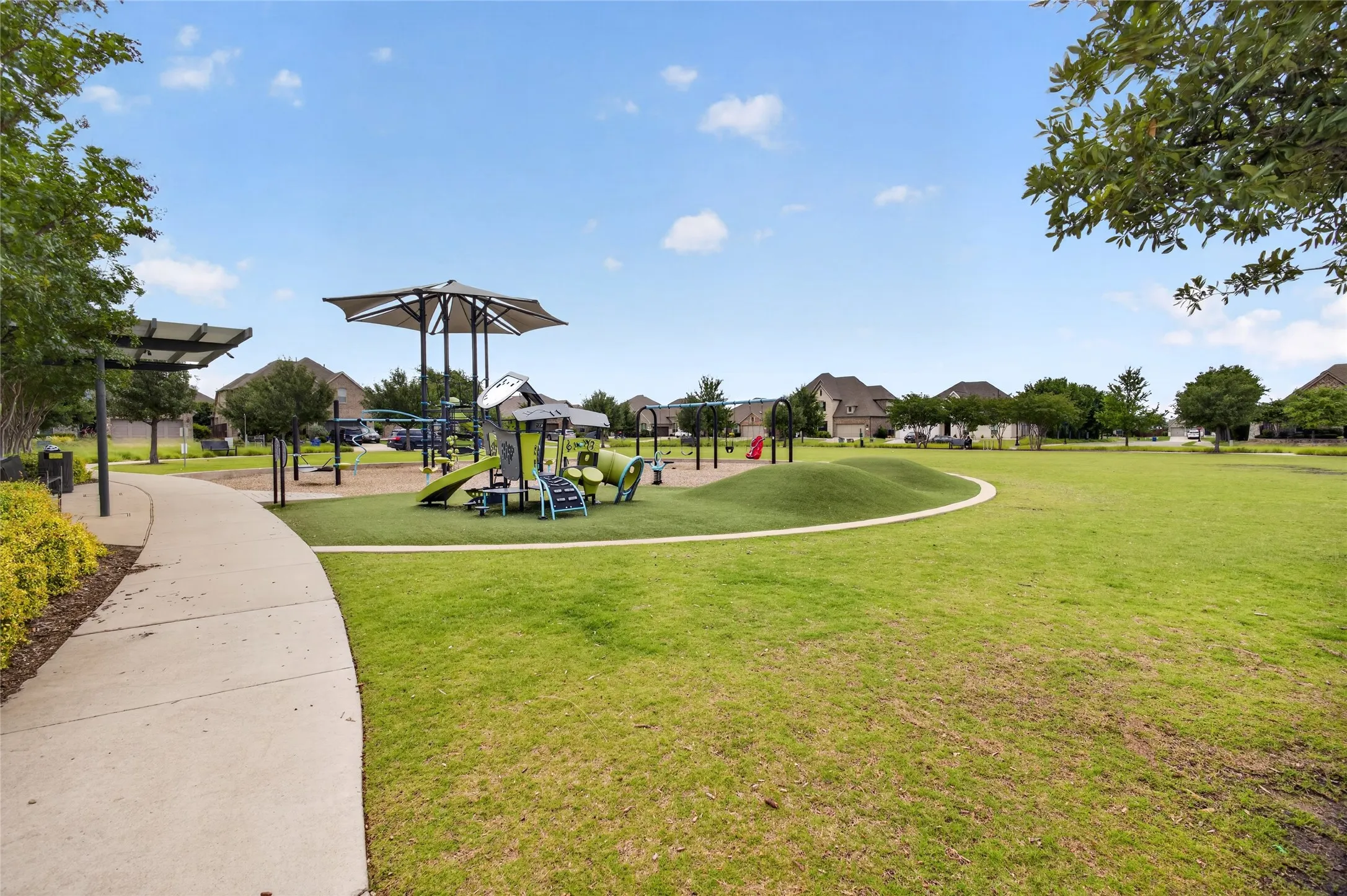 Community park featuring a modern playground structure with shade canopies, a curvilinear concrete pathway, and expansive green spaces