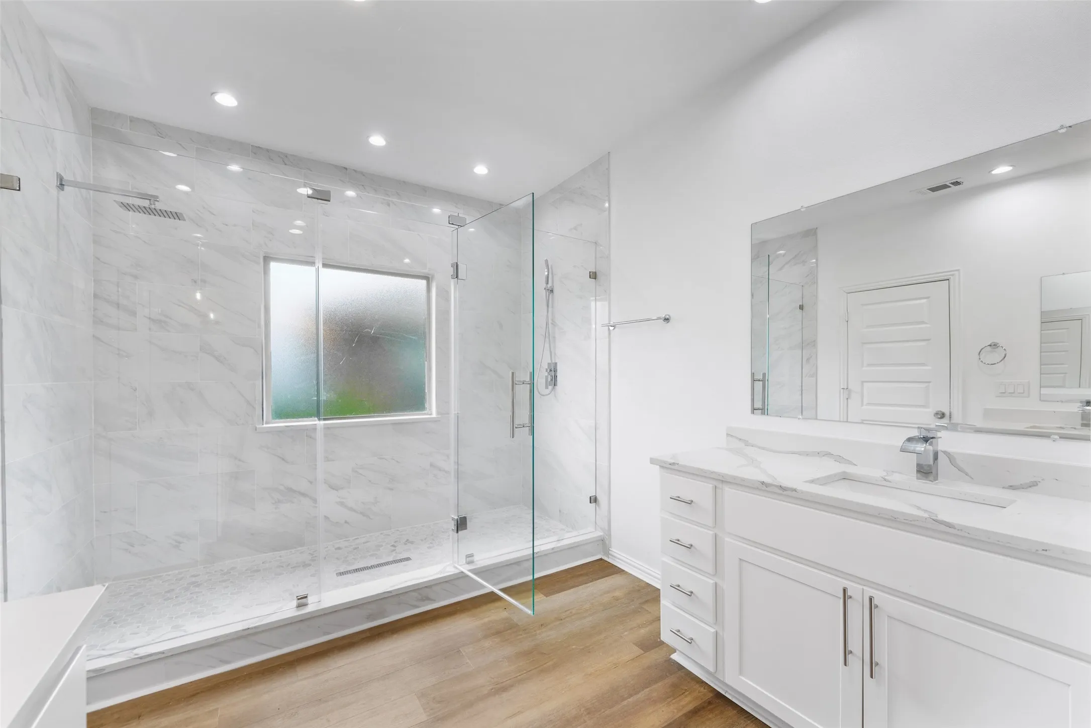 Spacious bathroom featuring a frameless glass shower enclosure with rain shower head, marble-patterned tile, a privacy window, a single vanity with stone countertop, and wood-finish flooring