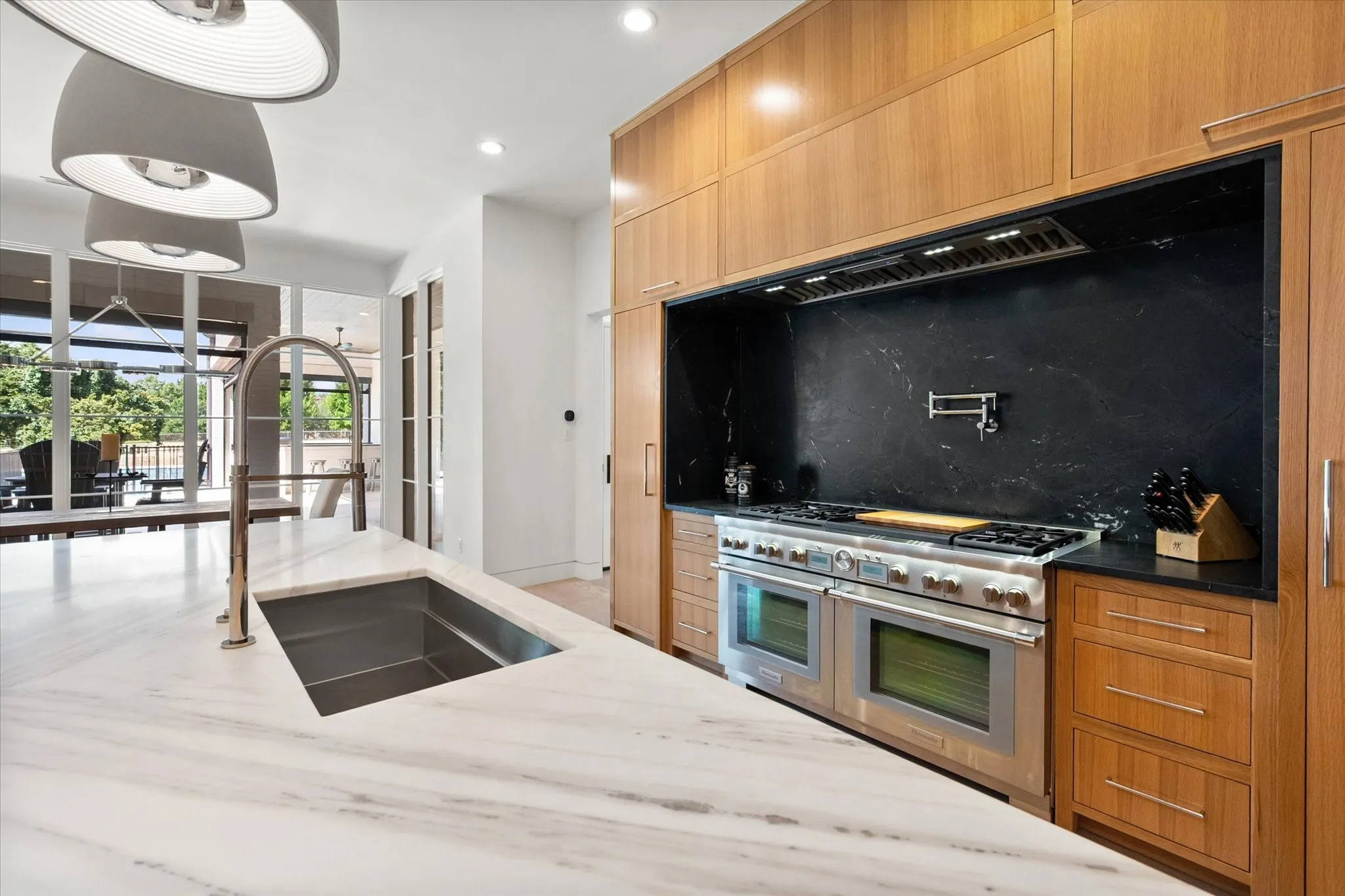 The Gourmet kitchen featuring a large island with an undermount stainless steel sink and gooseneck faucet, a professional-grade Thermador range with double ovens, a dark stone backsplash with a pot filler, wood cabinetry, and recessed lighting