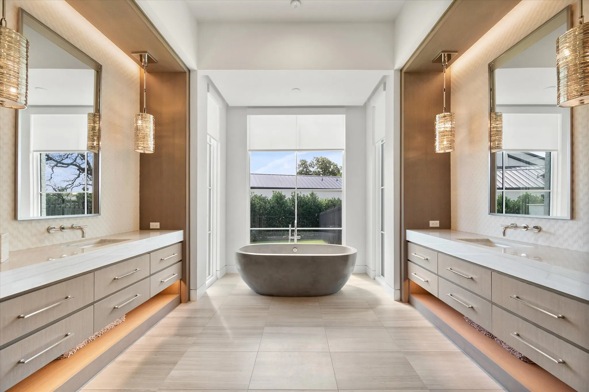 Freestanding soaking tub centered under a large window, flanked by dual floating vanities featuring under-cabinet lighting and stone countertops