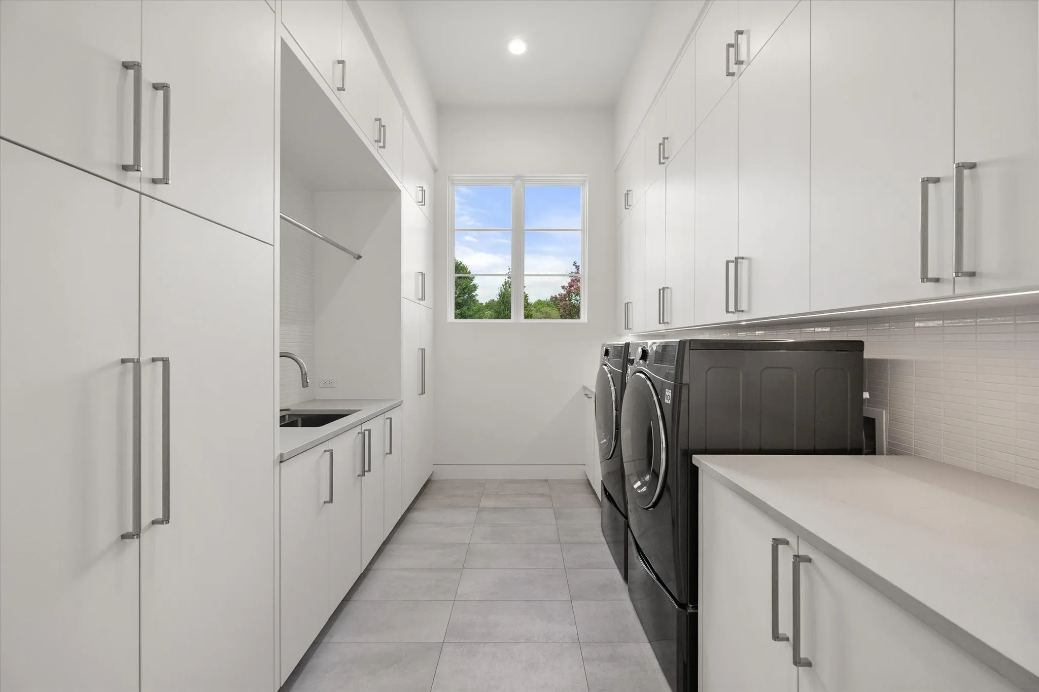 Dedicated laundry room featuring extensive white cabinetry, a built-in sink with a gooseneck faucet, light gray countertops, and neutral tile flooring