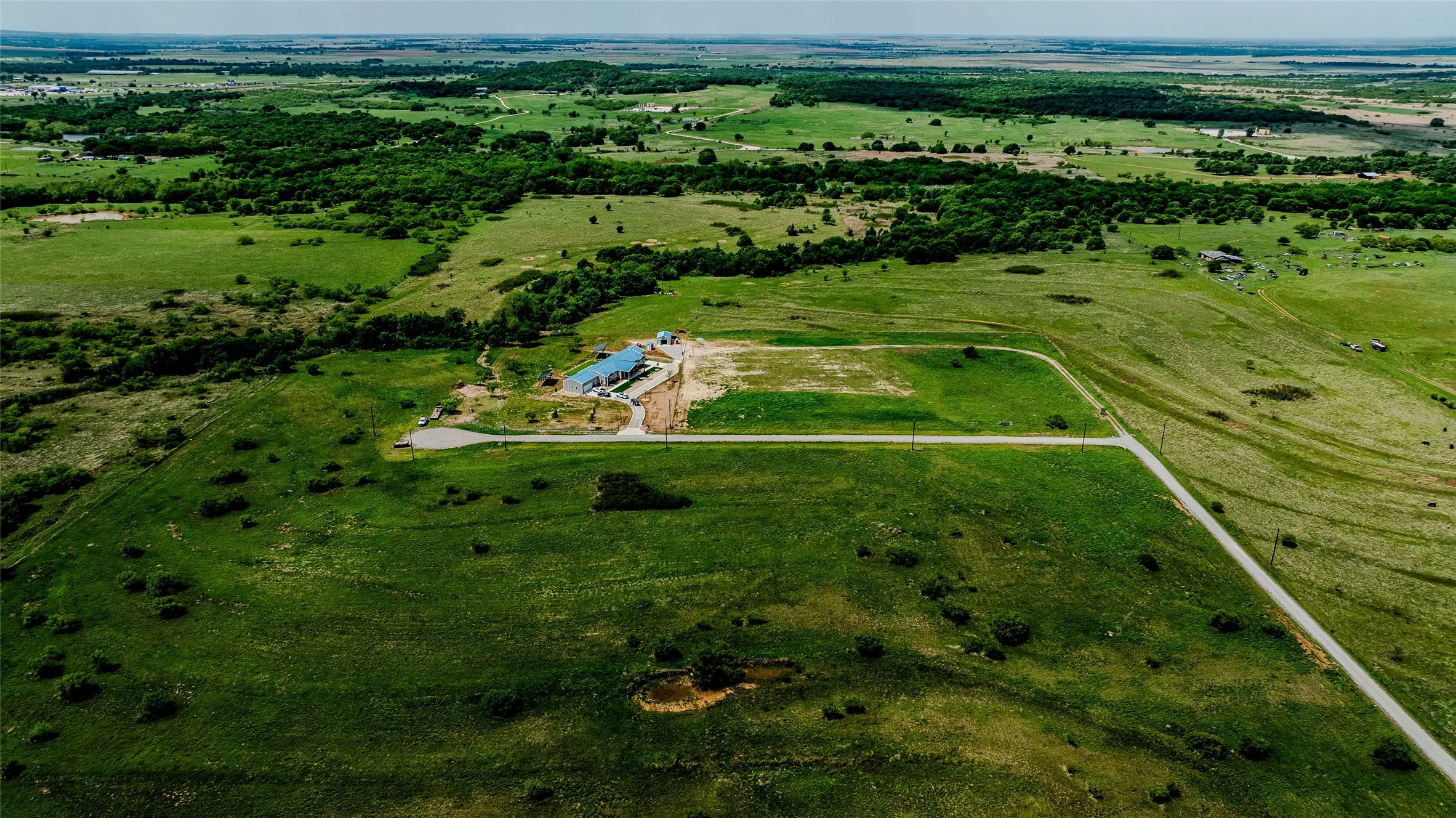 Expansive green acreage featuring a single-story structure with a blue roof, surrounded by open fields and distant tree lines
