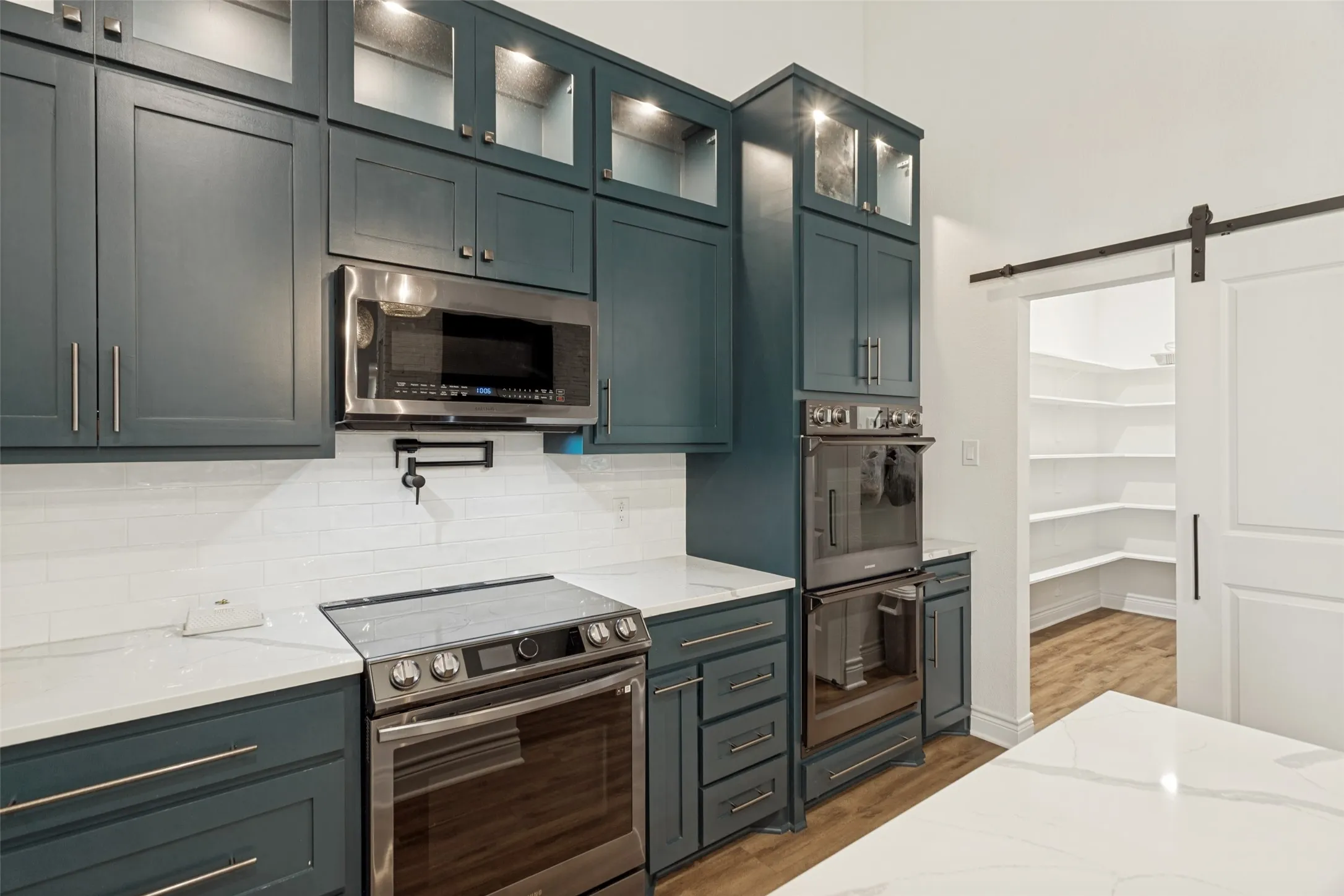 View of kitchen appliances (including 3 ovens!), electric cooktop, and pot filler.  Also a view into the spacious walk in pantry to store all food items and kitchen equipment with attractive barn door