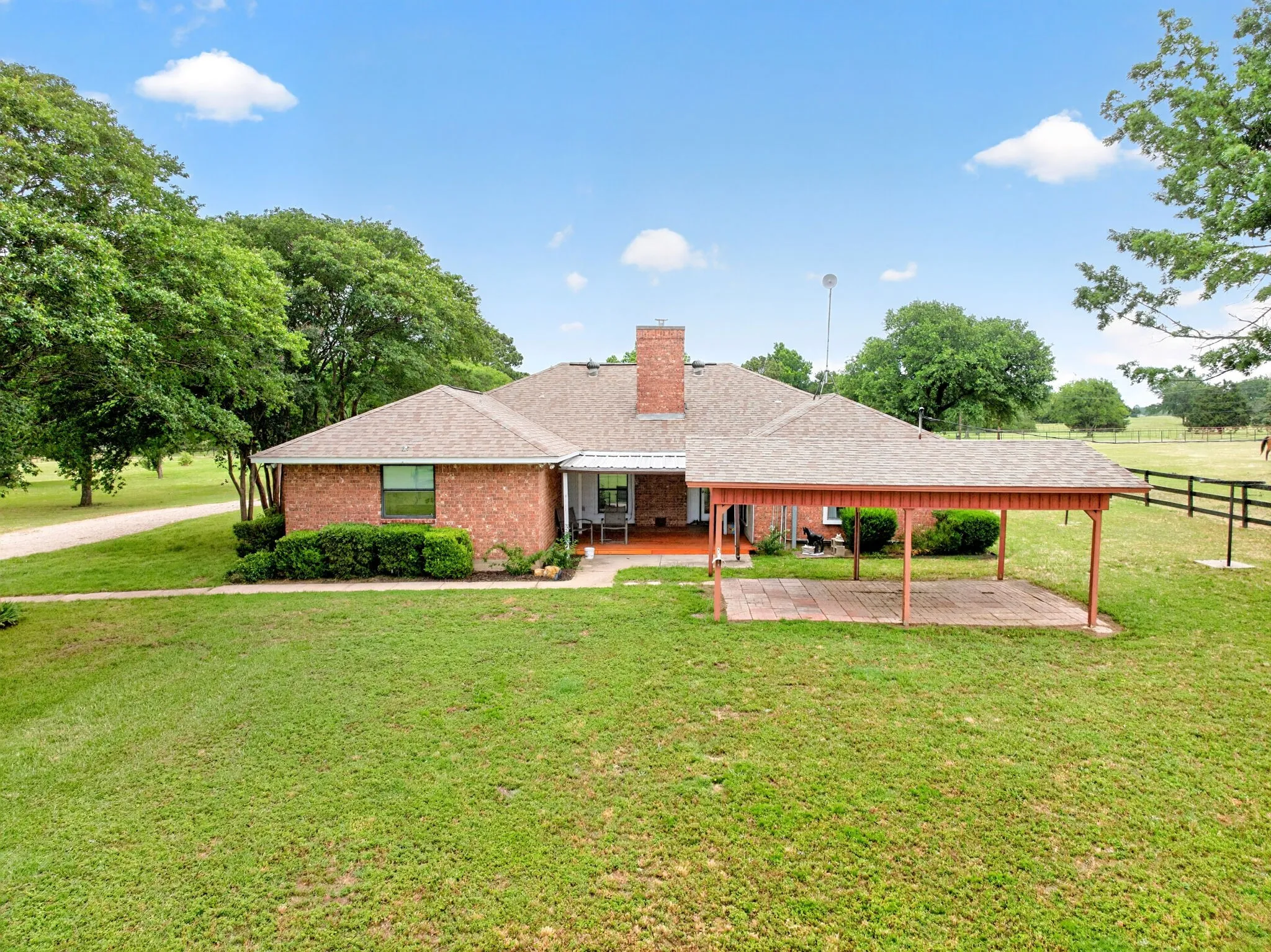 Brick exterior residence featuring a covered patio, extensive green lawn, mature trees, and an additional covered outdoor area with a paved surface
