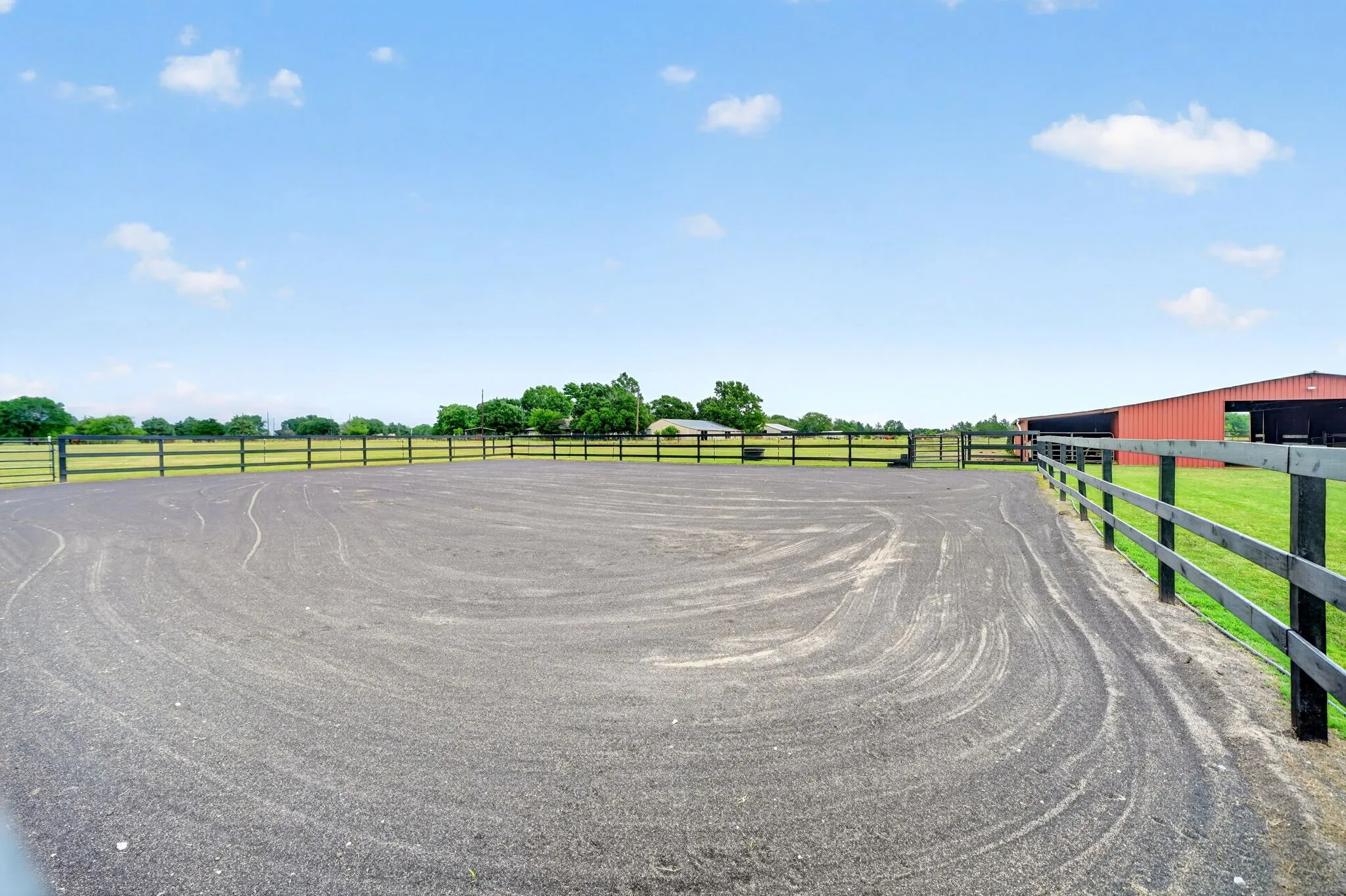 Expansive riding arena featuring a dark aggregate surface, surrounded by black post-and-rail fencing