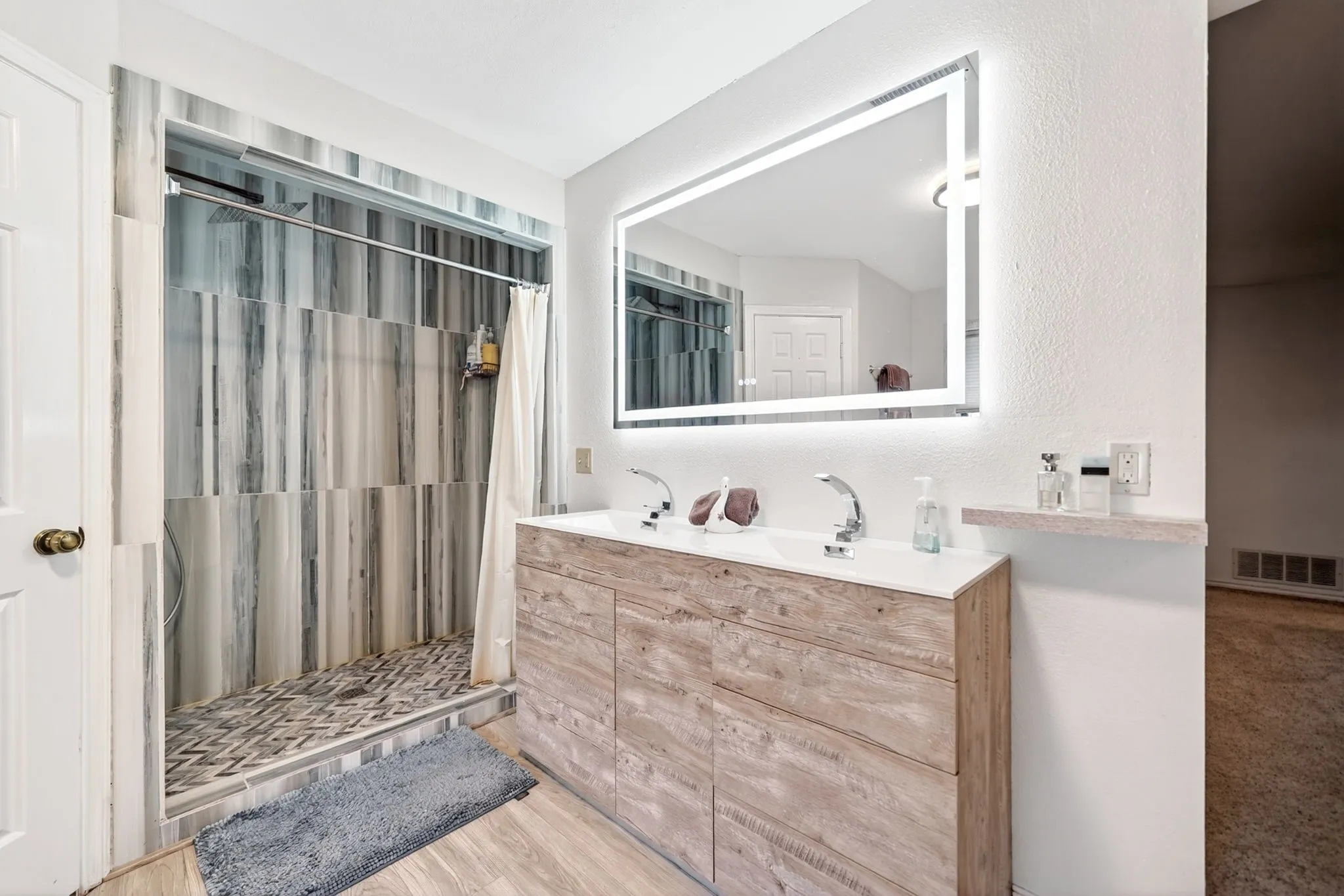 Bathroom with wood-finish flooring, a dual-sink vanity featuring a white countertop and chrome fixtures, and an illuminated rectangular mirror