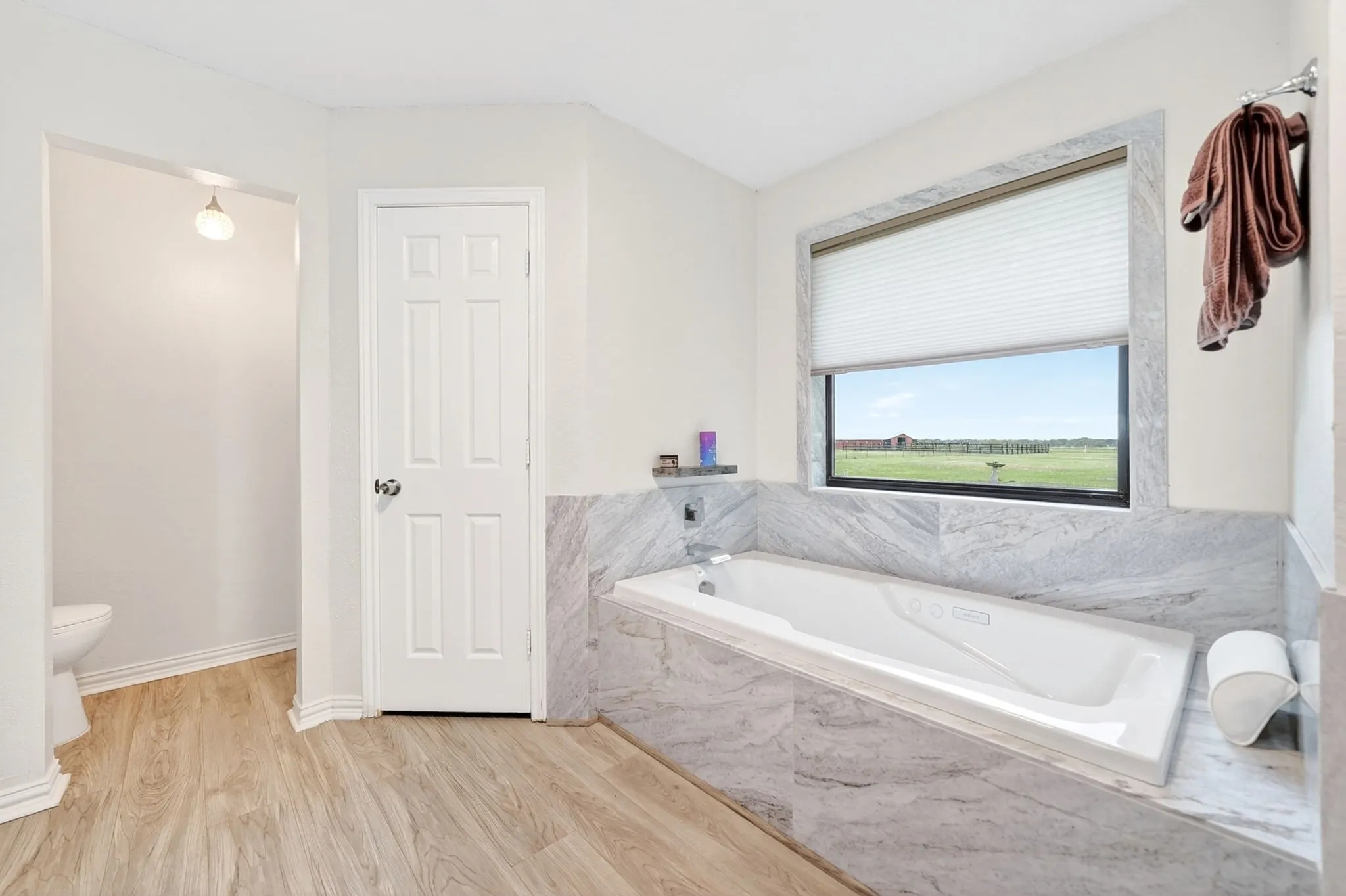 Soaking tub with stone-finish surround, large window with expansive outdoor scenery, wood-finish flooring, and white paneled door