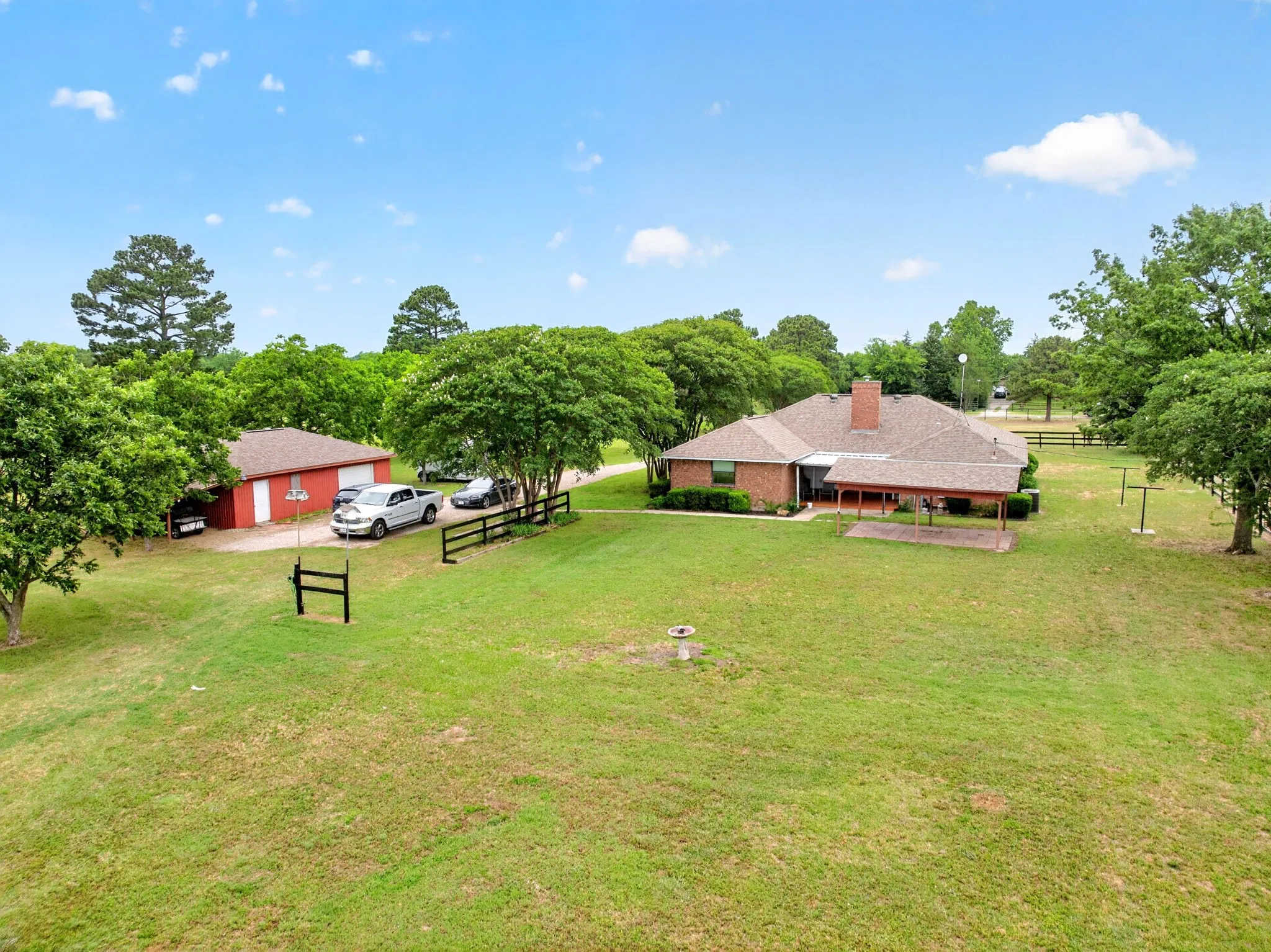 Expansive property featuring a brick residence, detached red barn with covered parking, lush green lawn, mature trees, and perimeter fencing