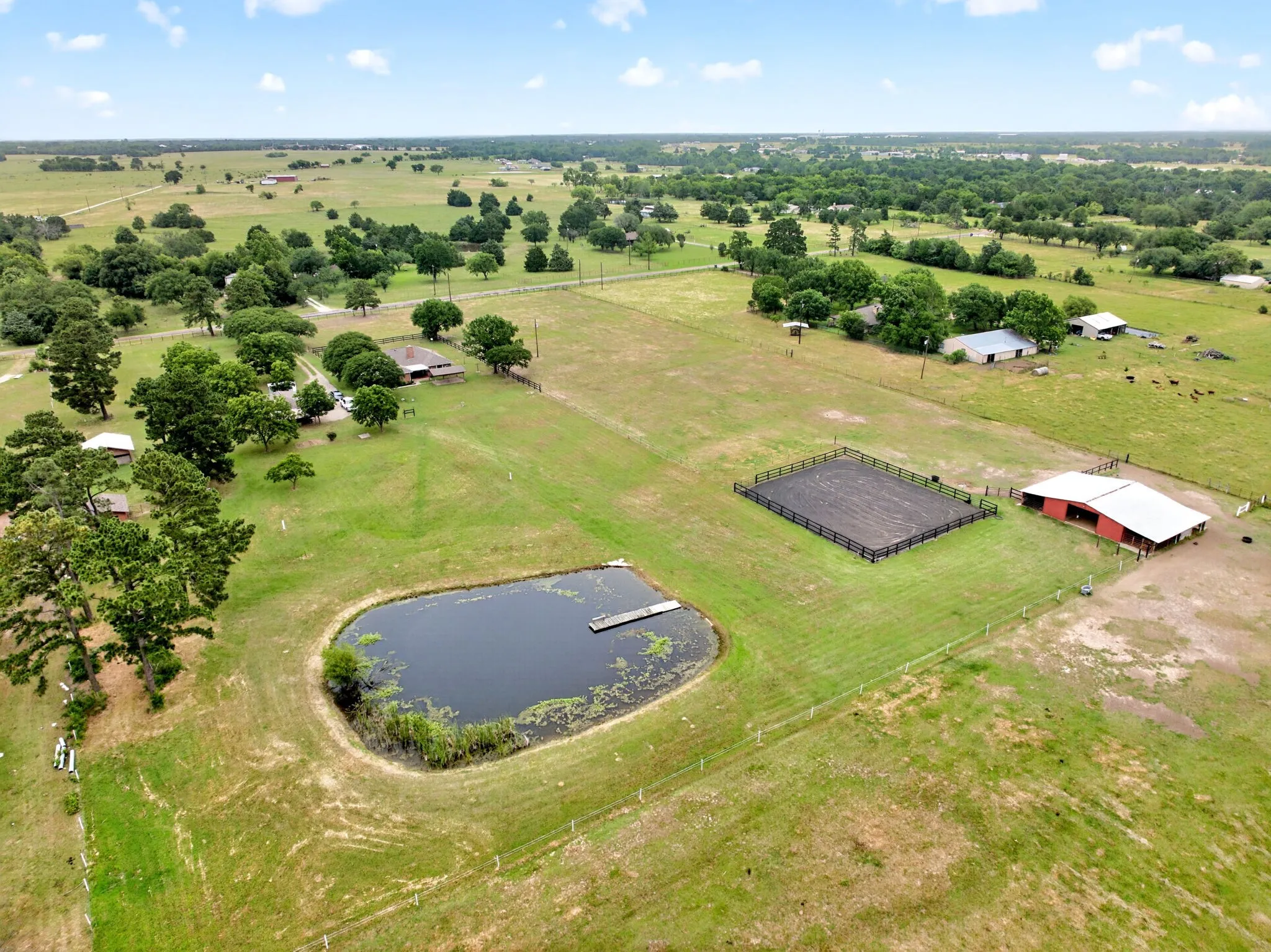 Expansive rural property featuring a private pond with a small dock, a red barn with an attached covered area, and a fenced riding arena