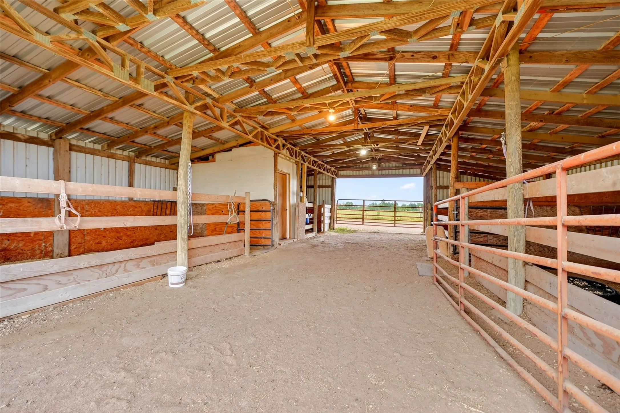 Spacious barn interior featuring exposed wooden rafters, a corrugated metal roof, multiple stalls with wooden fencing, and a metal gate opening to an exterior pasture