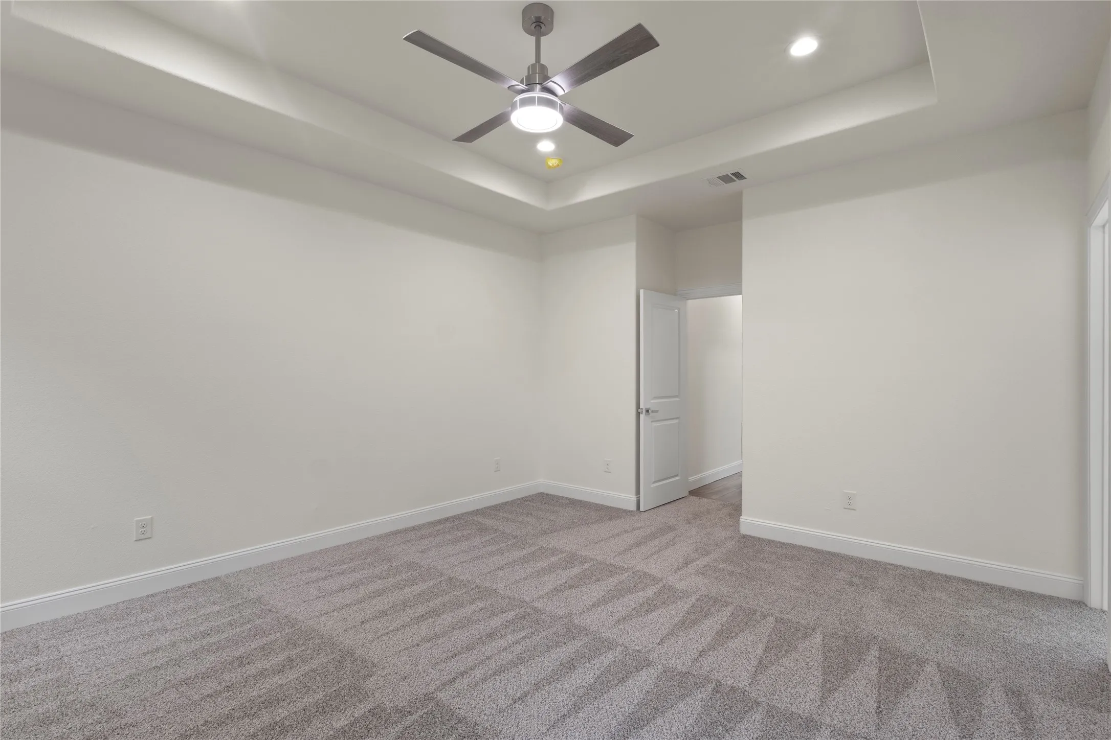 Spacious room featuring a tray ceiling with recessed lighting and a ceiling fan, light gray patterned carpet, white baseboards, and a white interior door