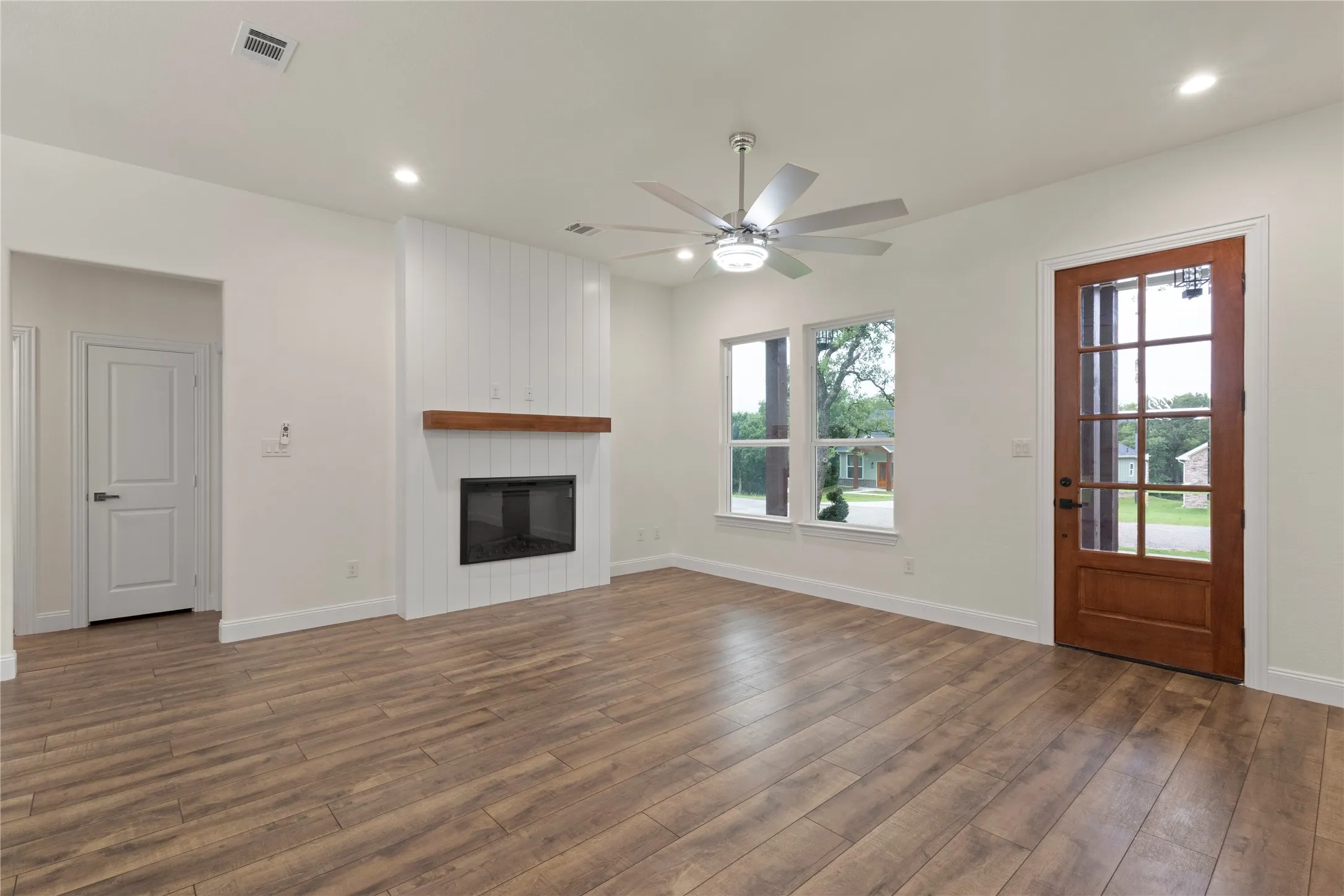 Spacious living area featuring wood-finish flooring, a contemporary fireplace with a white shiplap surround and wooden mantel, and a ceiling fan with integrated lighting