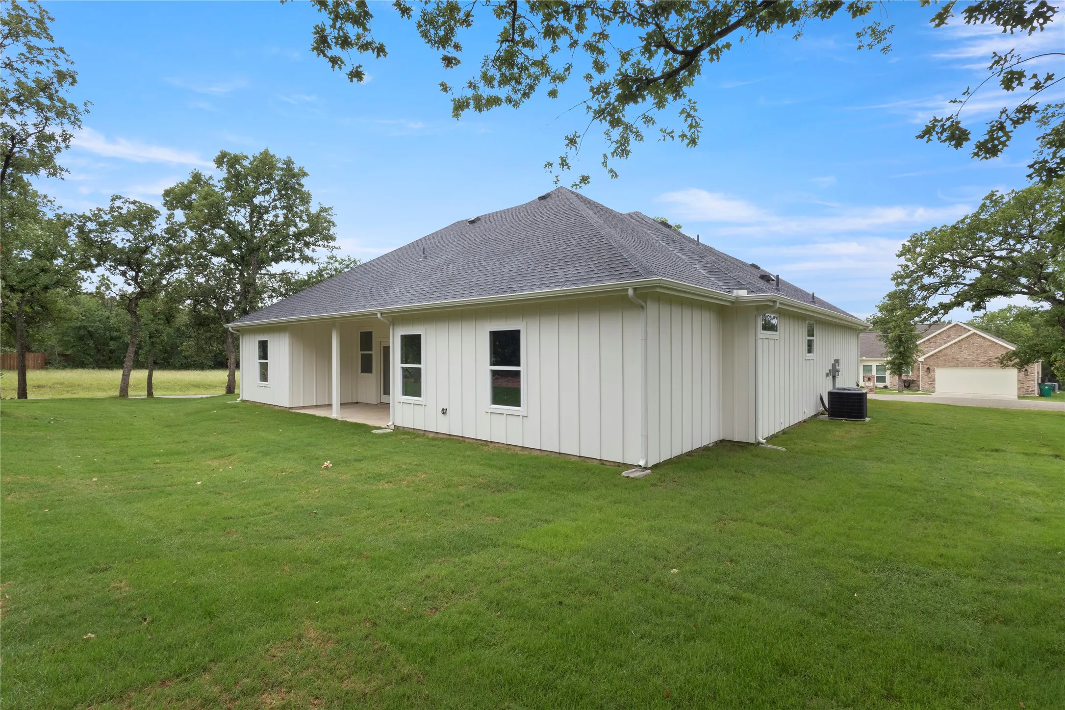 Expansive green lawn surrounds this property featuring white vertical siding, a dark shingle roof, and a covered concrete patio