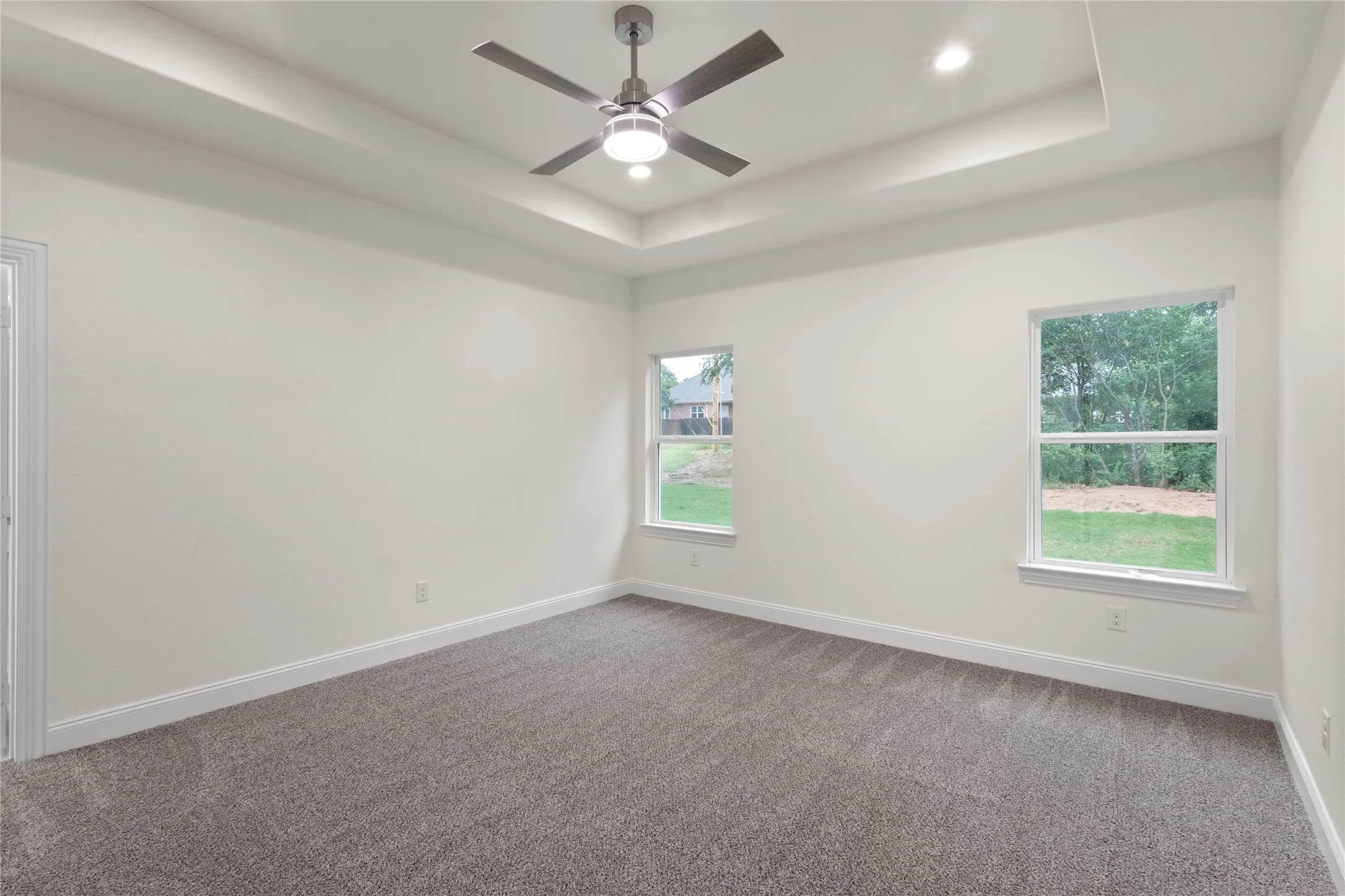 Spacious room featuring a tray ceiling with recessed lighting and a ceiling fan, plush gray carpeting, white baseboards, and two windows