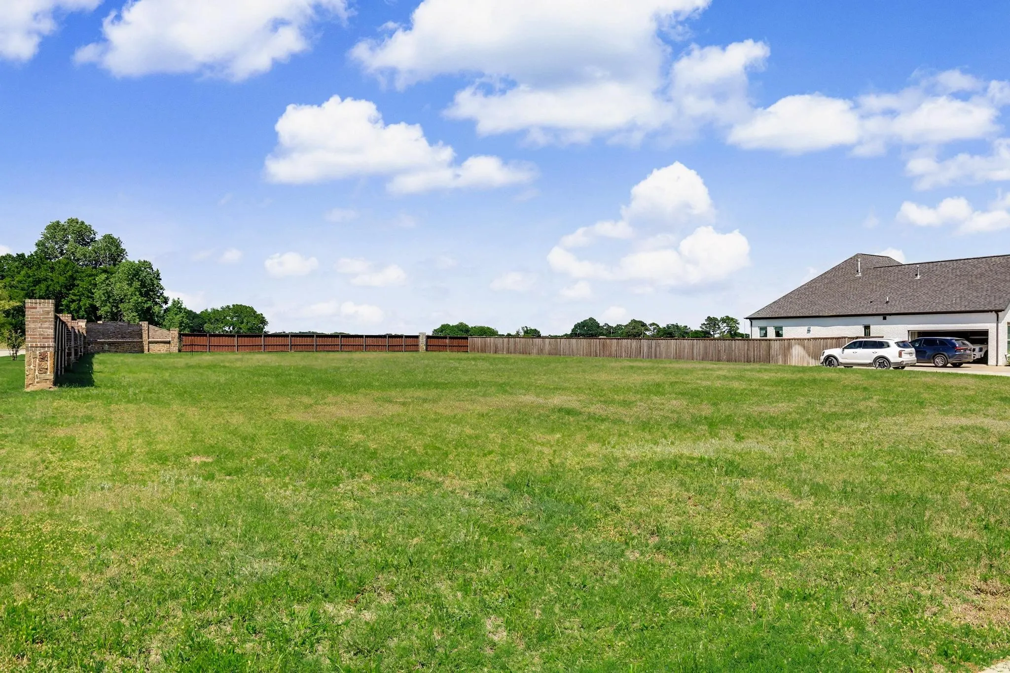 Expansive grassy lot with a wooden privacy fence, brick fence pillars, and mature perimeter trees