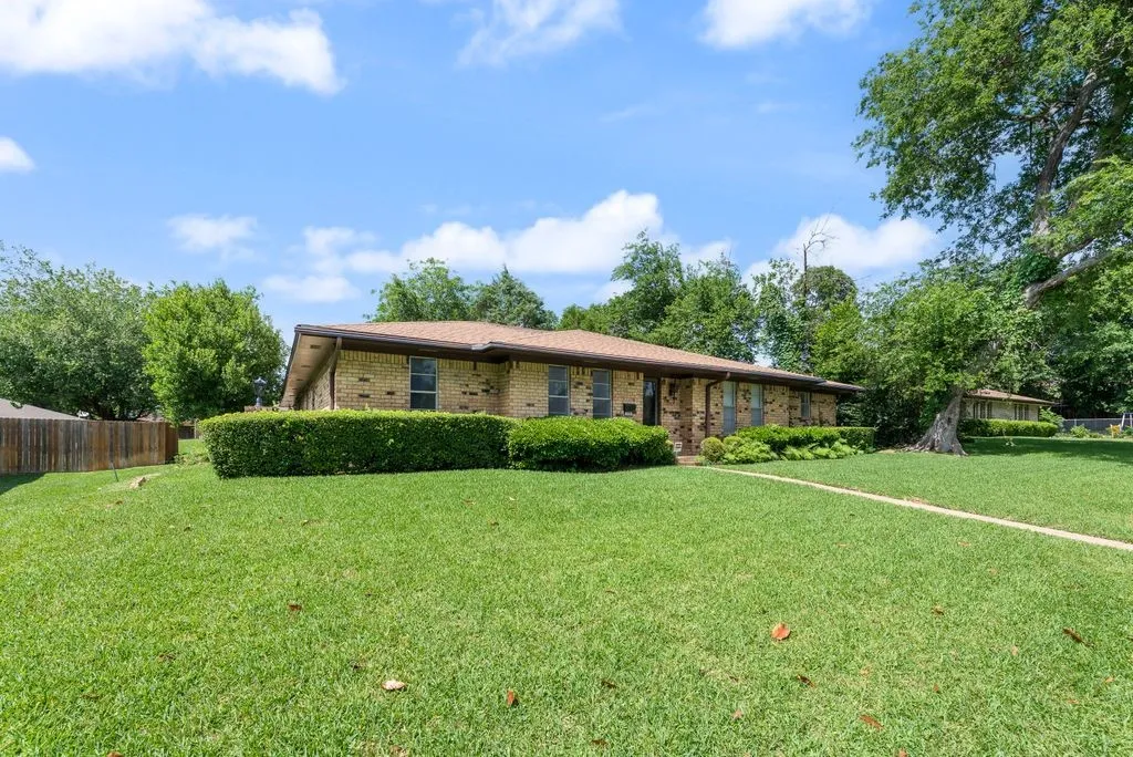 Brick exterior home featuring a brown shingle roof, well-maintained front lawn, mature perimeter trees, and manicured shrubbery