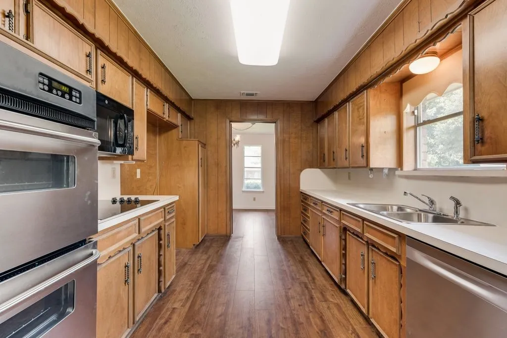 Galley kitchen featuring wood-finish flooring, wood paneling, wood cabinetry, and stainless steel appliances including a built-in oven and microwave